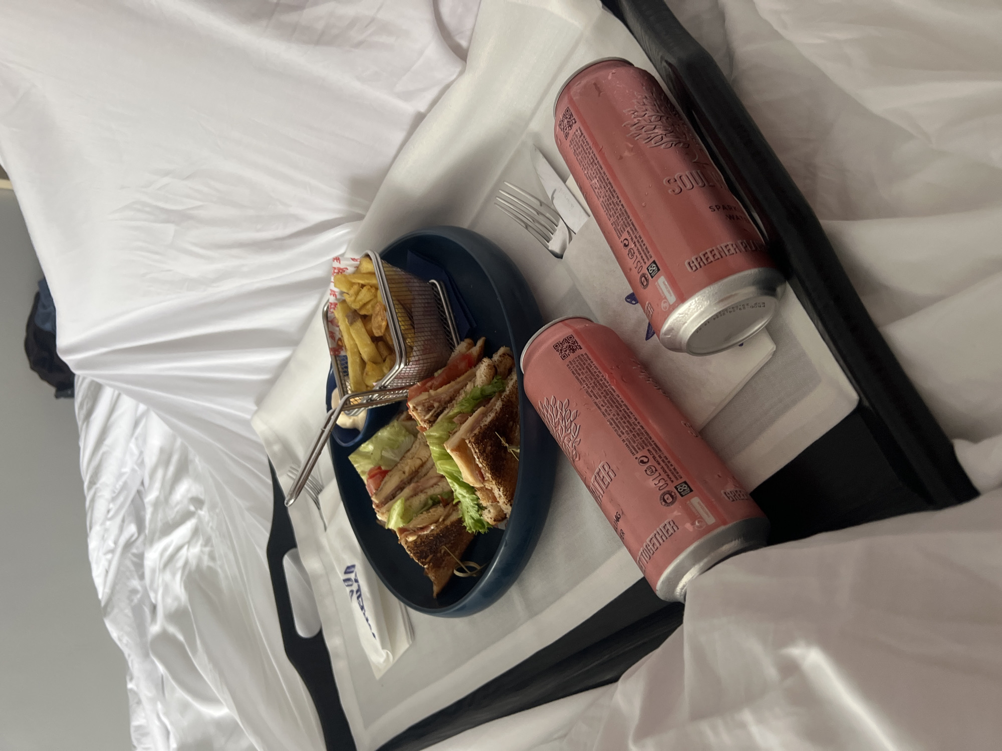 On a white bed in Santa Eulària des Riu, Spain, sits a black tray with a meal. The tray contains a blue plate holding a cut club sandwich and a metal basket of french fries. Cutlery rests beside the plate. Two pink cans of "Soul Water" sparkling water are also on the tray. A white napkin with blue writing lies near the plate. The tray has black handles. The bed's white sheets are crumpled.