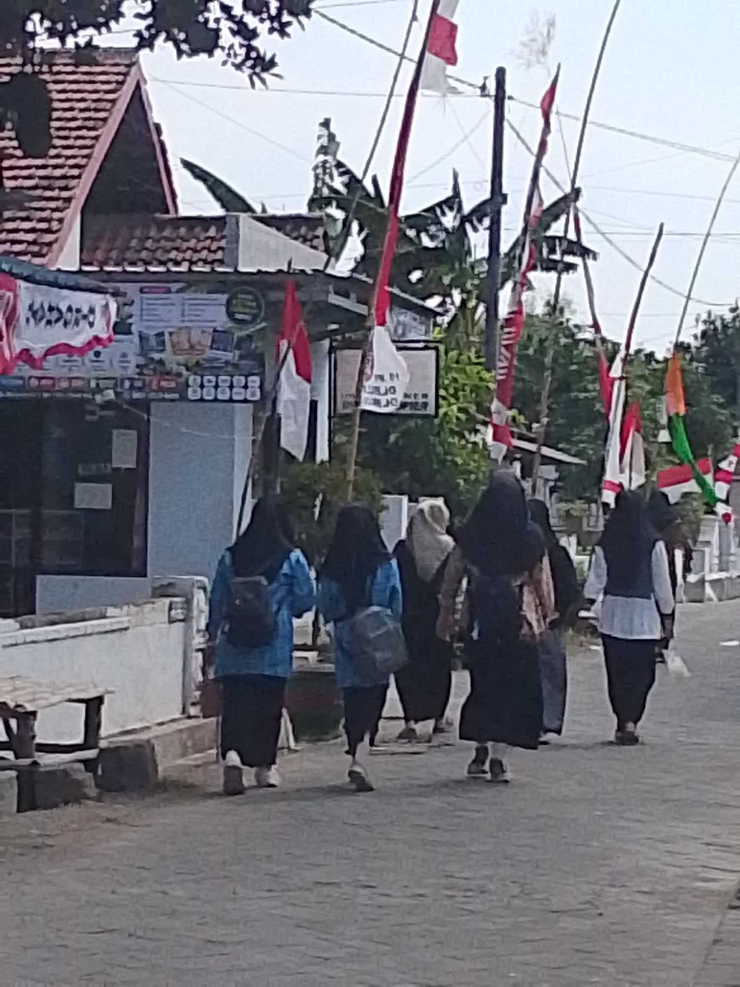 The image depicts a street scene in Bulurejo, Indonesia, with several individuals walking along a paved road. The foreground shows the lower bodies of five people, all wearing modest clothing, including headscarves. The people are walking away from the camera, suggesting a procession or a casual stroll. The individuals are dressed in various colors and styles, with some wearing dark-colored long skirts and tops, while others have on blue jackets and black skirts. Backpacks are visible on some of them.
In the background, a building with a tiled roof and signage is visible on the left side of the street. Red and white flags are prominently displayed on poles along the street, some appearing to be Indonesian flags, indicating a possible national celebration or event. Trees and foliage are present behind the building and along the street, suggesting a semi-urban or rural setting. The sky is overcast. The overall impression is of a community event or daily life in a Indonesian town.