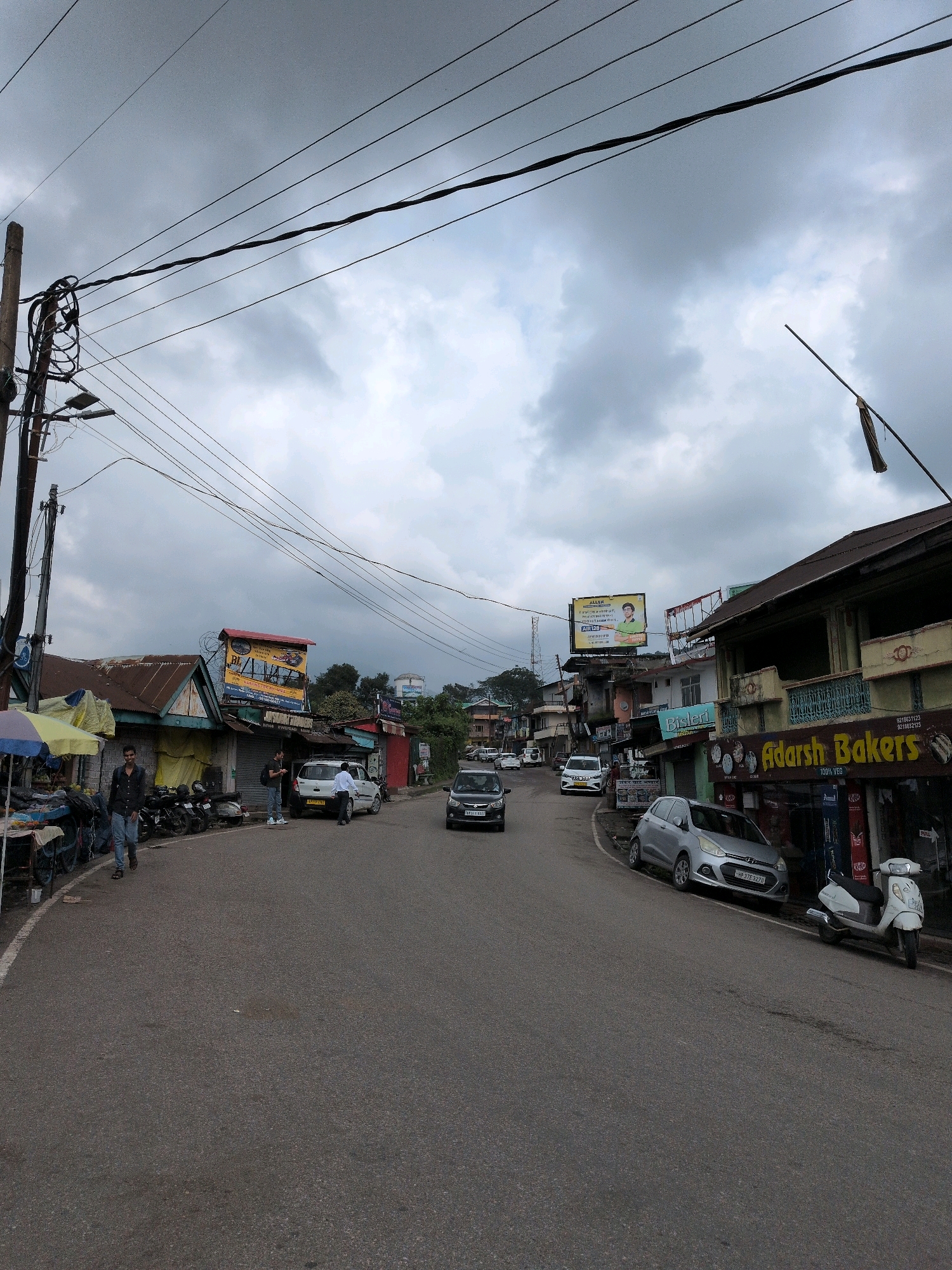 The image depicts a street scene in Baijnath, India, under a cloudy sky. The road is paved with asphalt and has a white line marking the edge of the lane on the left. On the left side of the road, there are several small shops with corrugated iron roofs. One shop has an umbrella providing shade, and a person is standing next to a cart laden with goods. Further down the street, there are parked motorcycles and a white car.
On the right side of the street, a row of buildings houses businesses. A prominent sign reads "Adarsh Bakers." Other signs indicate businesses like "Amul." A silver car is parked along the curb in front of the buildings, and a white scooter is parked behind it.
In the background, more buildings and trees are visible. Overhead, a complex network of electrical wires is strung between utility poles. A large billboard advertising "ALLEN" is visible on top of one of the buildings. The overall impression is a busy commercial street in a town.