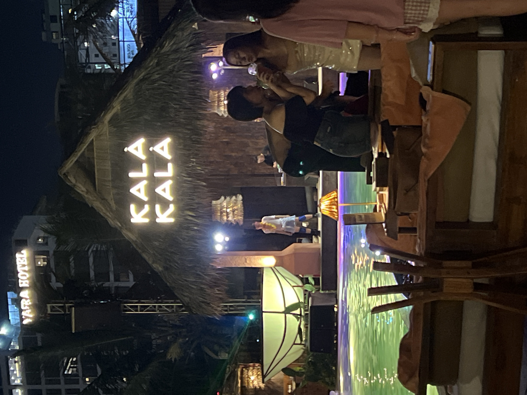 The image captures a nighttime scene at the "KALA KALA" establishment, identified as being in the city of Đà Nẵng, Vietnam. Above the thatched-roof structure, the sign for "YARA HOTEL" is visible on a building. In the foreground, people are seated at wooden tables and chairs, with orange towels draped over them, positioned near a brightly lit swimming pool. In the background, a performer in a white shirt and lei stands on a stage. Two women are at a table, with one appearing to hand a drink to the other, who is wearing a sparkly dress. The scene is illuminated by artificial lights, creating a vibrant and festive atmosphere.