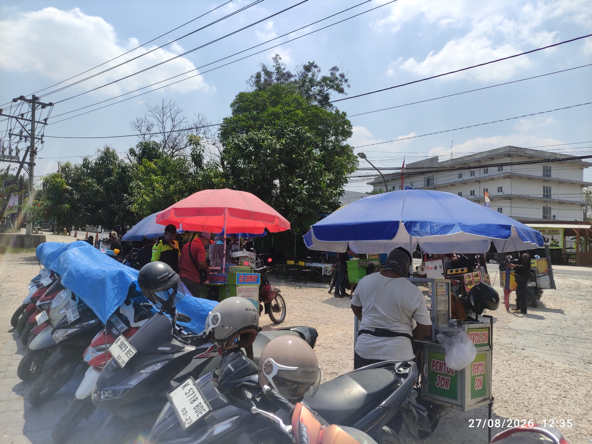 A sunny, outdoor scene in Tamanrejo, Indonesia, depicts a row of parked motorcycles covered with blue tarps. Two vendors operate under colorful umbrellas, one red and one blue and white, beside portable food carts. The red umbrella vendor, wearing a red shirt, interacts with customers. Under the blue and white umbrella, a vendor in a white t-shirt prepares food. The background features a multi-story building, trees, power lines, and a streetlamp. A timestamp in the lower right corner reads "27/08/2026 12:35". Several helmets are visible on the parked motorcycles.