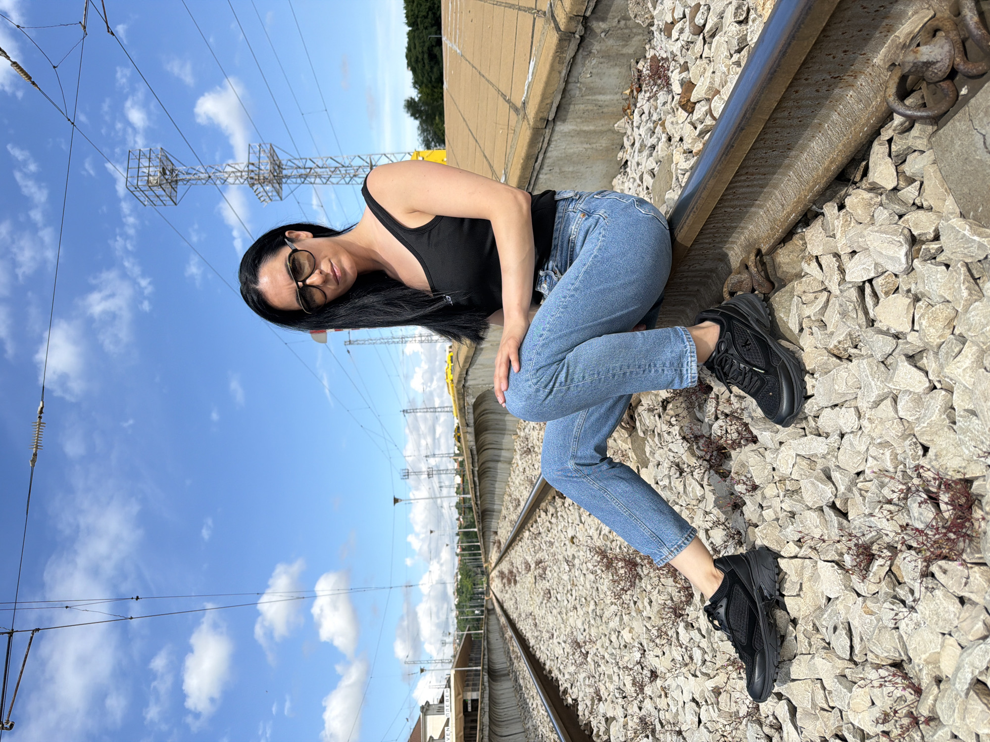 A woman with long black hair and sunglasses is sitting on the gravel next to a railway track. She is wearing a black tank top and blue jeans. She is looking at the camera. In the background, there is a train station with a building and a sign that reads "PLEVEN". There are also power lines and a tower in the background. The sky is blue with some clouds. The scene is located in the city of Pleven, Bulgaria.