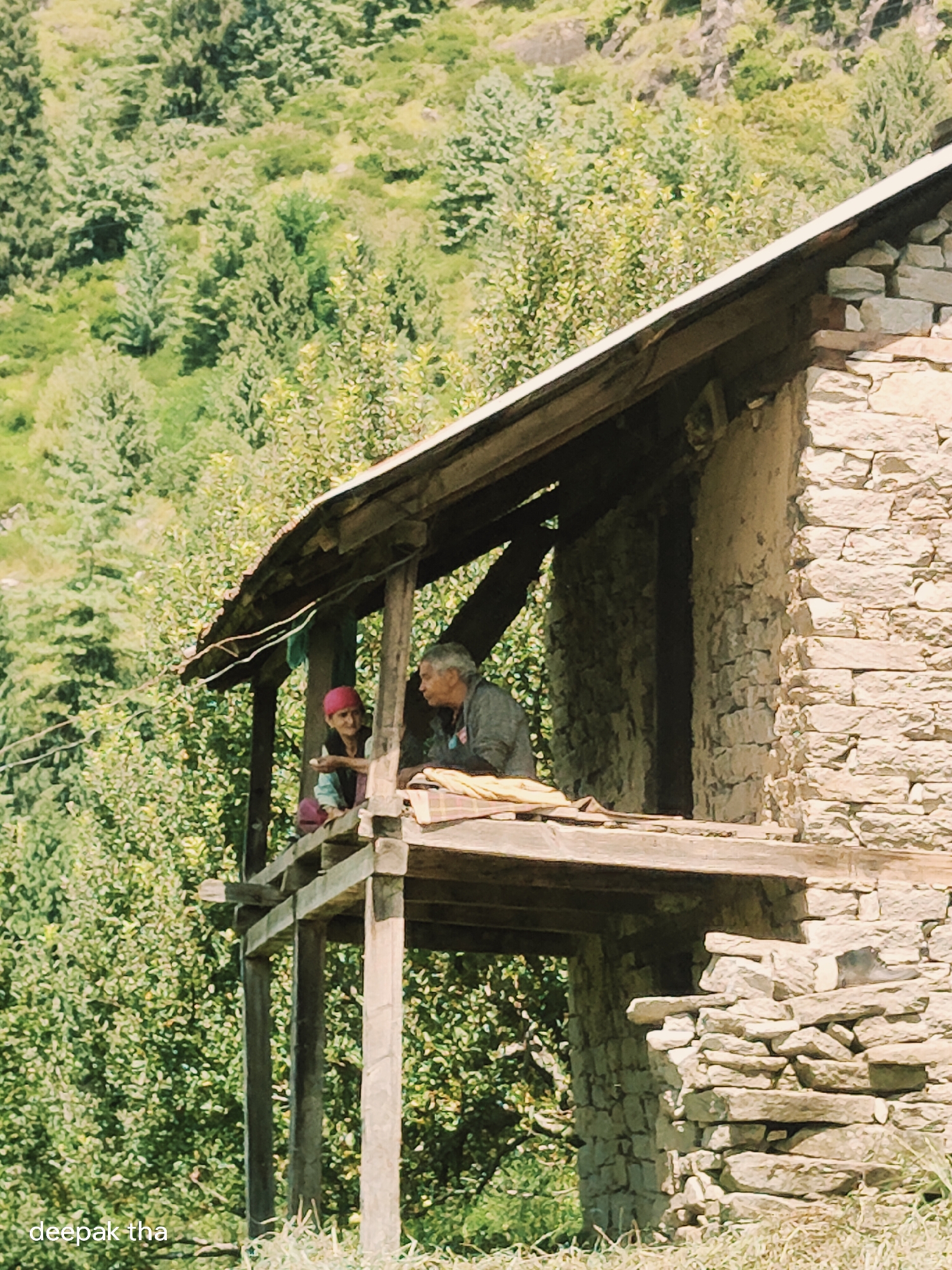 An outdoor, mid-shot photograph captures a scene in Vashisht, India, featuring two individuals near a stone structure. The building, constructed with rough-hewn stones, has a wooden balcony supported by thick beams. The roof of the balcony is made of corrugated metal and wooden planks.
On the balcony, an older individual with gray hair, wearing a dark-colored jacket, is seated and looking towards the left. To their left, a younger person, wearing a light blue shirt and a red headscarf, is also seated and appears to be offering something to the older person. They are holding a small white object in their hand.
The background is a lush, green hillside covered with dense foliage and trees, typical of the mountainous terrain of Himachal Pradesh. The lighting suggests it is daytime, with soft shadows indicating a slightly overcast or diffused light. The overall impression is one of a quiet, rural setting. The lower left corner of the image contains the text "deepak tha".