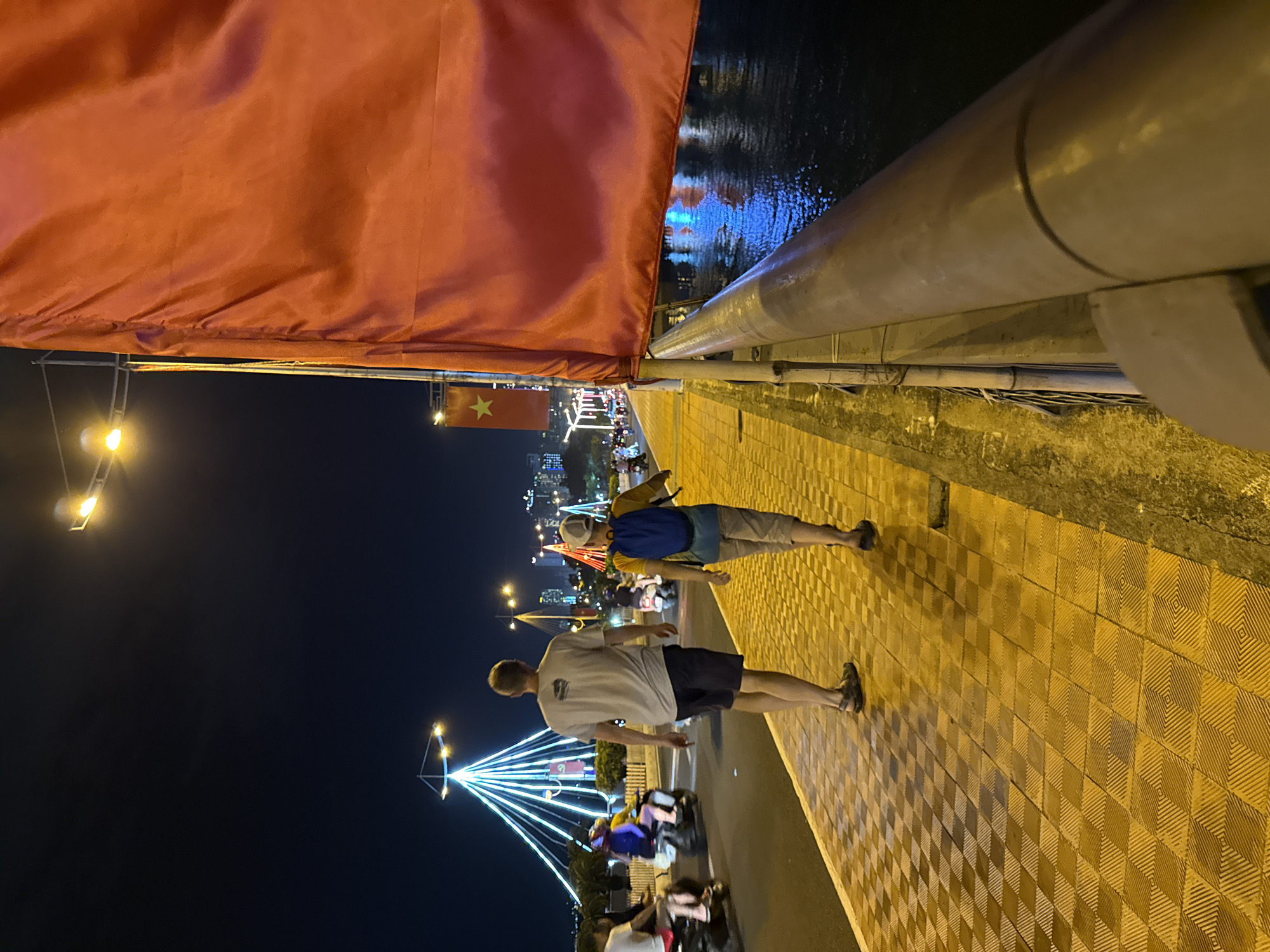 At night, two individuals are walking on a pedestrian path along a road in Nha Trang, Vietnam. The path is paved with yellow tiles in a diamond pattern. The road is wet, likely from recent rain, and reflects the lights of the city and decorative lighting. In the background, a large red flag with a yellow star, characteristic of the Vietnamese flag, hangs from a pole and partially obstructs the view. Decorative lights in the shape of a pyramid are visible on the left, along with other illuminated structures and city buildings in the distance. Several motorbikes are seen on the road, some with riders. The sky is dark, indicating it is nighttime. The overall scene suggests a city scene at night, possibly during a celebration or event.