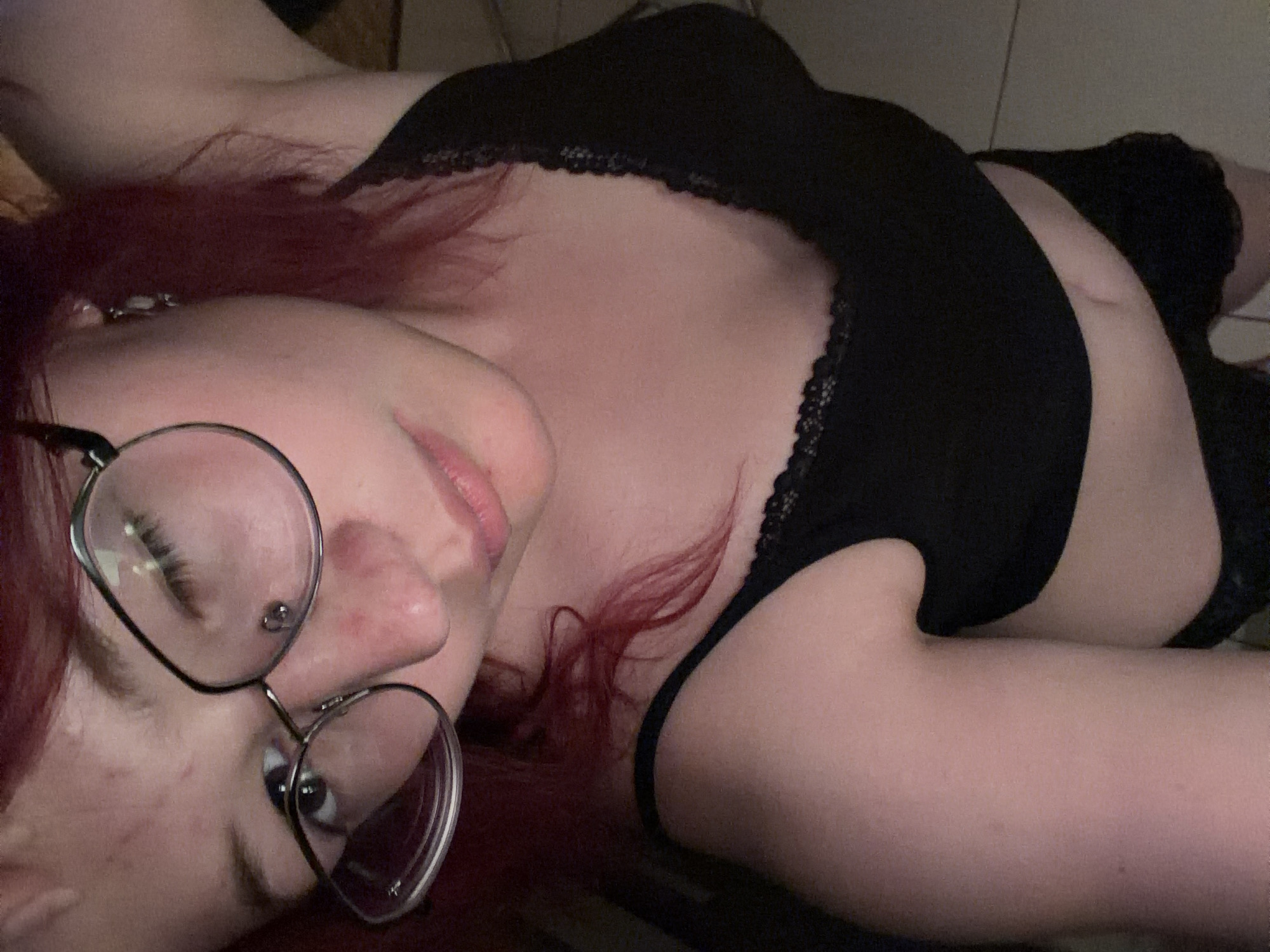 The image is a close-up, overhead selfie of a young woman with red hair and glasses. She is wearing a black tank top with lace trim. Her skin appears to have some redness and blemishes. The background is indistinct, appearing to be an indoor setting with tiled flooring. There are no discernible landmarks or environmental cues to suggest a specific location within Chelyabinsk, Russia. The image focuses tightly on the woman's face and upper body.