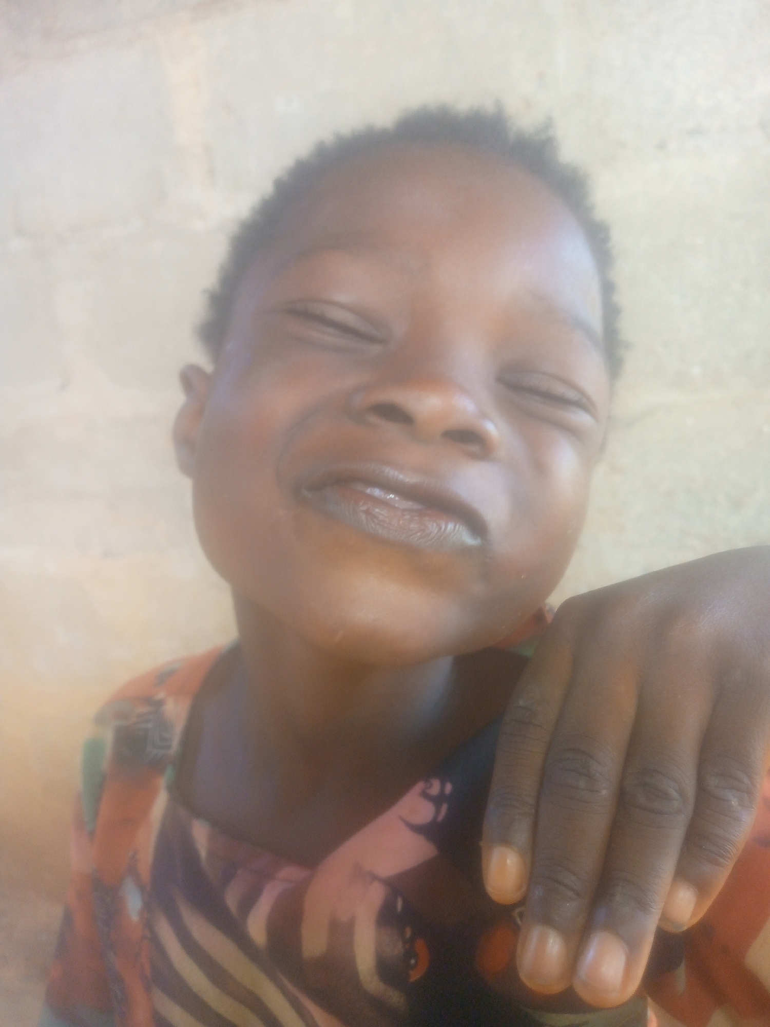 A close-up, slightly blurred portrait captures a young child with dark skin and short, dark, curly hair, likely in a joyful moment. The child's eyes are tightly closed or squinting, and a wide, happy smile is spread across their face, revealing teeth and glossy lips. The expression conveys strong happiness, amusement, or perhaps a reaction to bright light.
The child is wearing a patterned garment, possibly a shirt or dress, featuring a mix of warm colors such as orange, red, and black, with geometric or abstract designs. A hand, presumably the child's, rests on the right side of the frame, showing visible fingers and fingernails that appear lighter in color.
The background is a plain, light-colored wall or surface, possibly beige or light yellow, suggesting a simple and uncluttered setting. No specific location cues beyond a wall are discernible. The lighting appears to be natural, indicating it is daytime. There is no visible text in the image. The overall impression is one of genuine childhood joy.