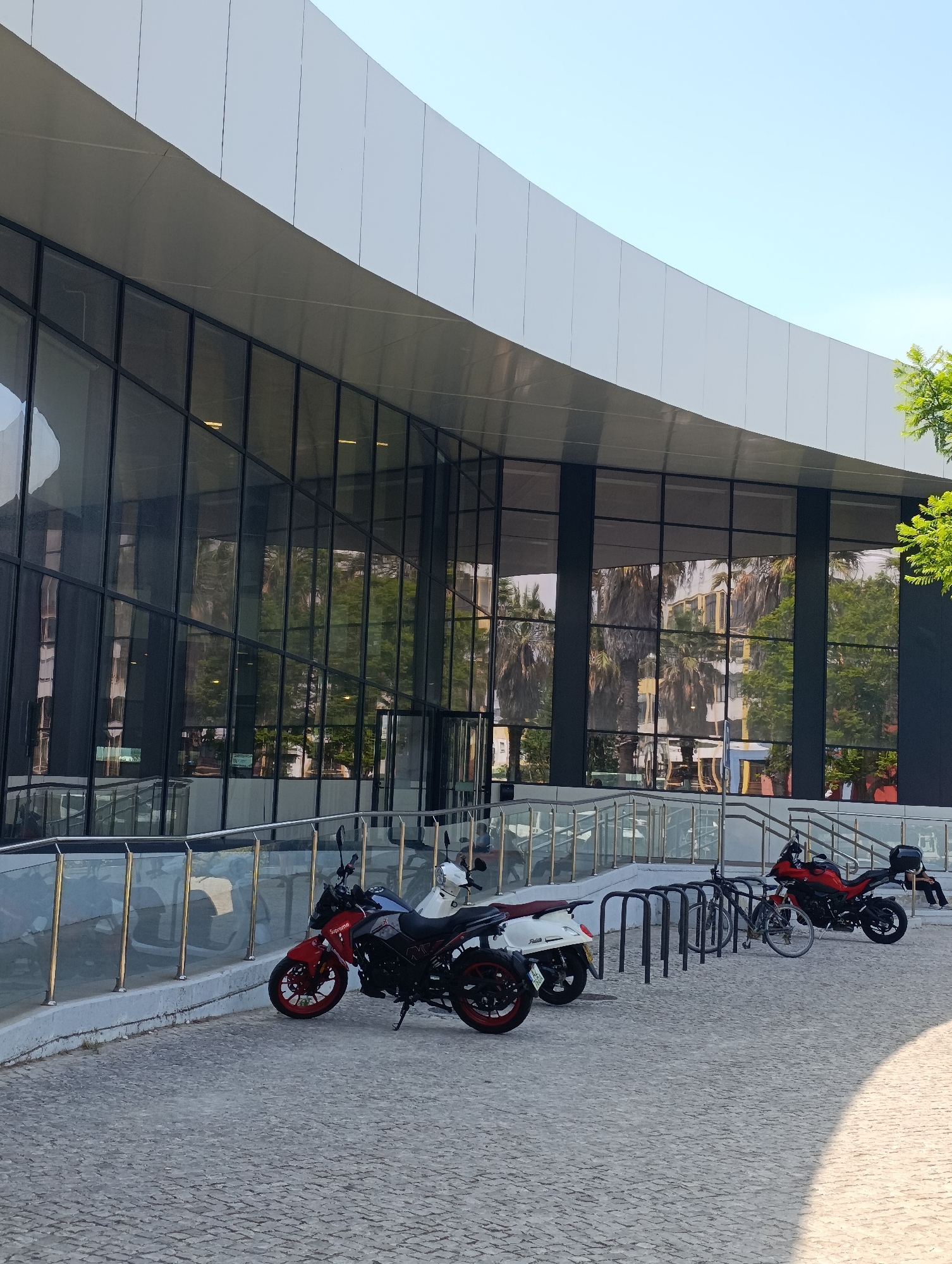 A modern building with a curved facade, featuring large glass windows, is situated in Setúbal, Portugal. The facade reflects palm trees and other foliage. In front of the building, on a cobblestone ground, are two motorcycles and two bicycles parked at a bike rack. One motorcycle is red and black, with red rims, and the other is white. One bicycle is silver and the other is darker colored. A metal railing runs along a raised pathway in front of the building. The sky is clear and blue.