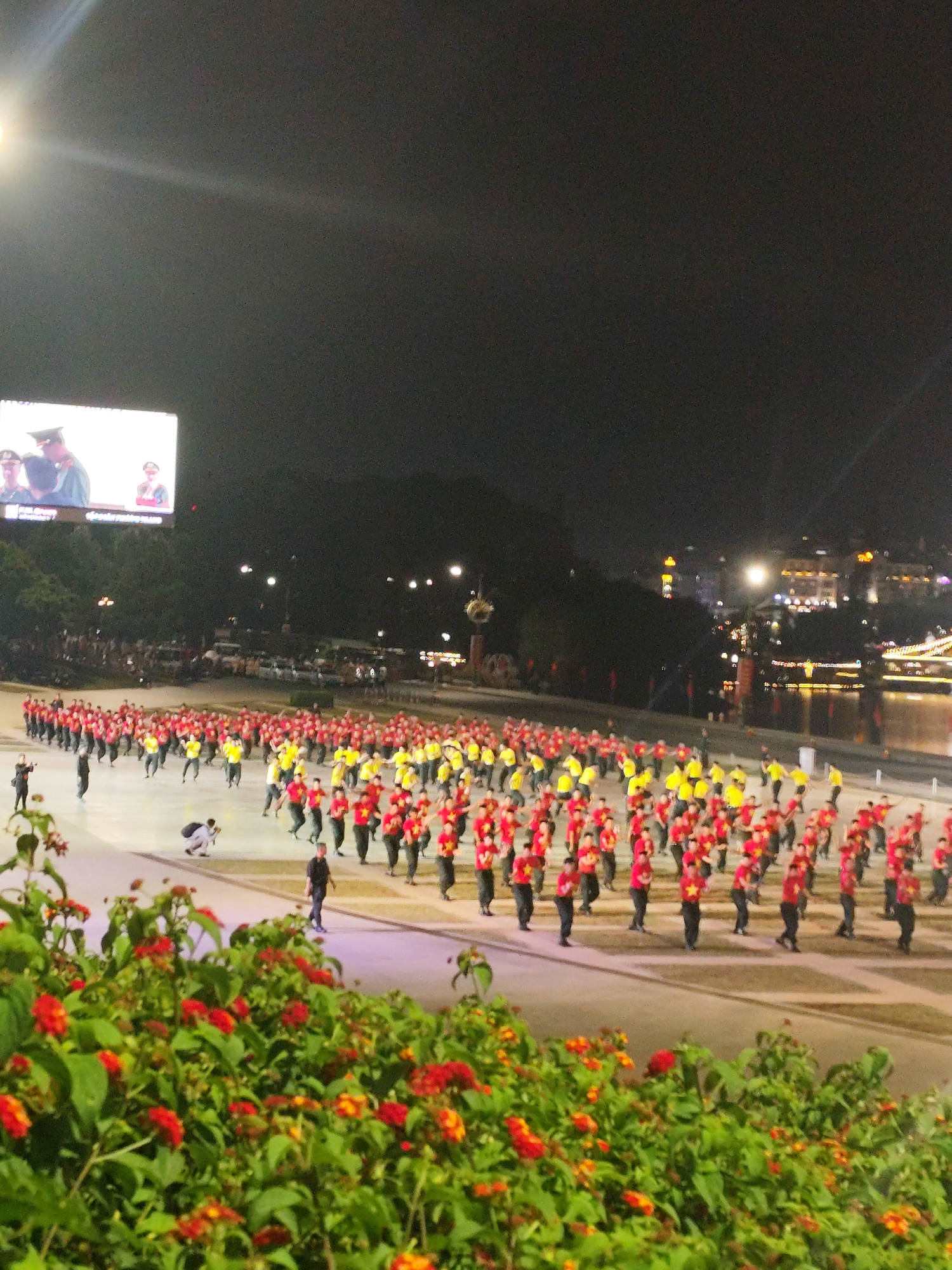 A large outdoor event is taking place at night in what appears to be Da Lat, Vietnam, with a crowd of people performing in unison. The scene is illuminated by bright lights, including a large screen displaying images of people in uniform. In the foreground, lush green foliage with vibrant red and orange flowers adds a natural element to the urban setting.
The primary subjects are the performers, a massive group of individuals dressed in bright yellow and red attire, spread across a wide paved area. They appear to be engaged in a synchronized dance or performance, with many in motion. Several individuals in black are also visible, some appearing to direct or observe the performers, while others are in the midst of the crowd. The background reveals a cityscape at night, with illuminated buildings and structures, suggesting an urban environment with water features, possibly a lake or river, given the reflections.
The activity is clearly a performance or ceremony of some kind, taking place after dark, as indicated by the artificial lighting and dark sky. The bright lights suggest it's an evening event. There are no discernible emotions on the faces of the performers due to the distance and resolution, but the organized nature of the event implies a sense of occasion and perhaps patriotism or community spirit.
Notable details include the large screen showing what seem to be military or official figures. The contrast between the high-tech screen and the mass of human performers, set against the backdrop of a city at night, creates a dynamic visual. The presence of the flowers in the foreground suggests the event is taking place in a park or public square. The weather appears clear, allowing for the large outdoor gathering. No visible text is present in the image, other than what might be on the large screen, which is not legible.