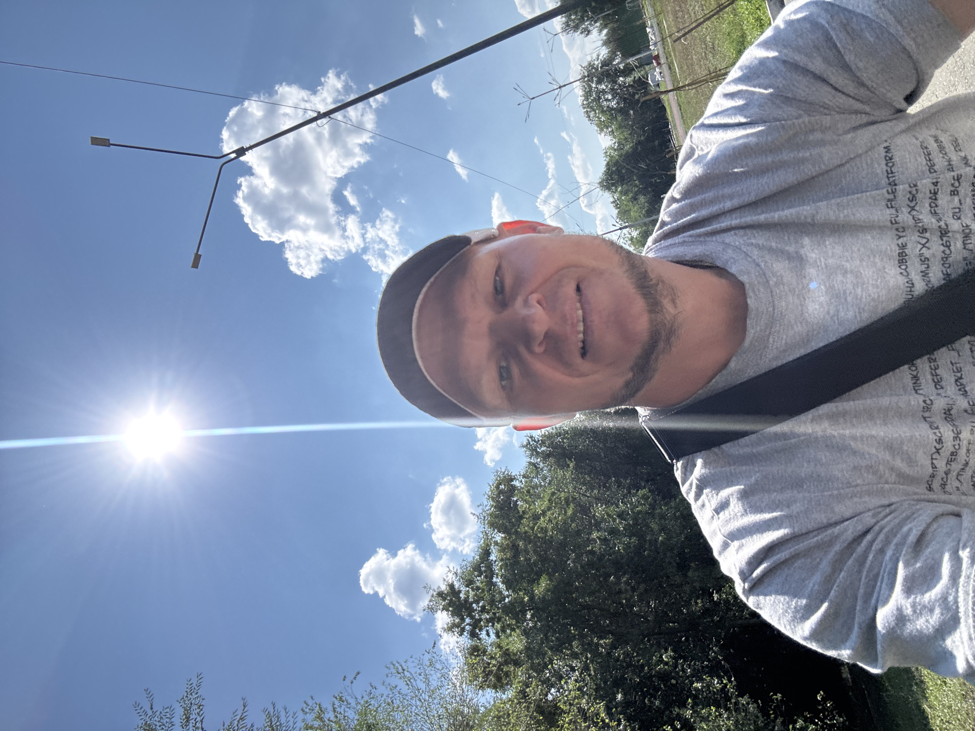 A man wearing a grey t-shirt and a baseball cap takes a selfie outdoors. The man is smiling and looking at the camera. In the background, there are trees, a blue sky with white clouds, a bright sun with lens flare, and streetlights. There is some text on the man's t-shirt, but it is not legible. The overall scene suggests a sunny day in an urban or suburban area.