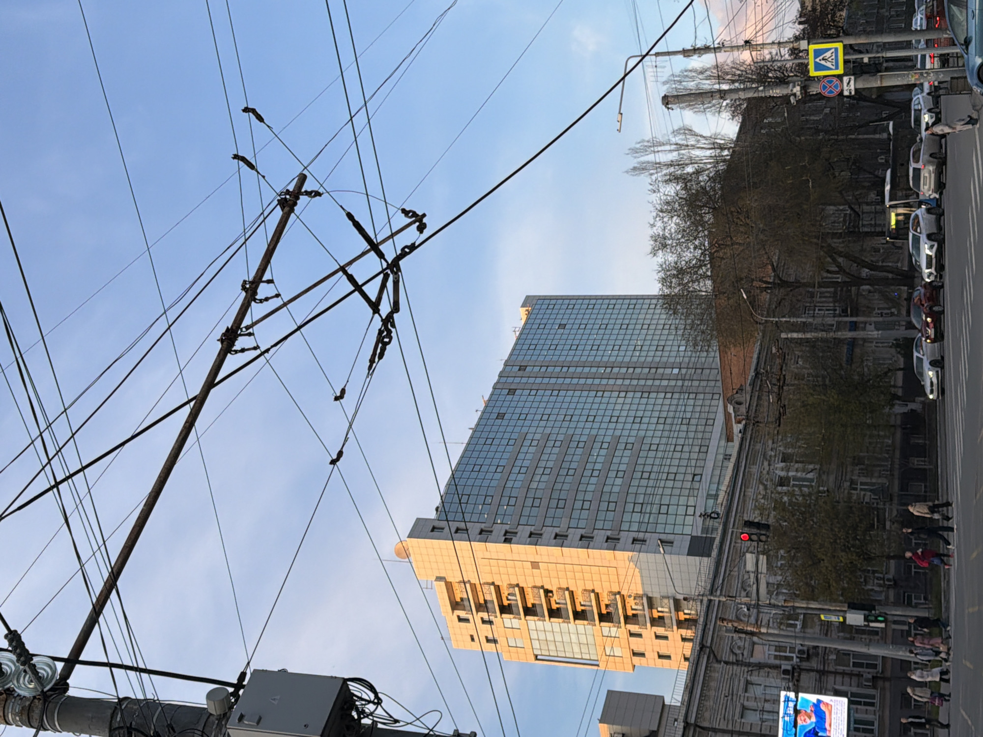 The image shows a street scene in Rostov-on-Don, Russia. A tall, modern, glass-facade building is visible, catching the golden light of either sunrise or sunset. In the foreground are overhead power lines and a utility pole. To the right of the building is an older, traditional building along a street. A pedestrian crosswalk is visible, with people crossing the street, indicating urban activity. Cars are parked alongside the street. The sky is clear and blue. A "pedestrian crossing" sign is present, as well as a red traffic light. A billboard is visible in the foreground.