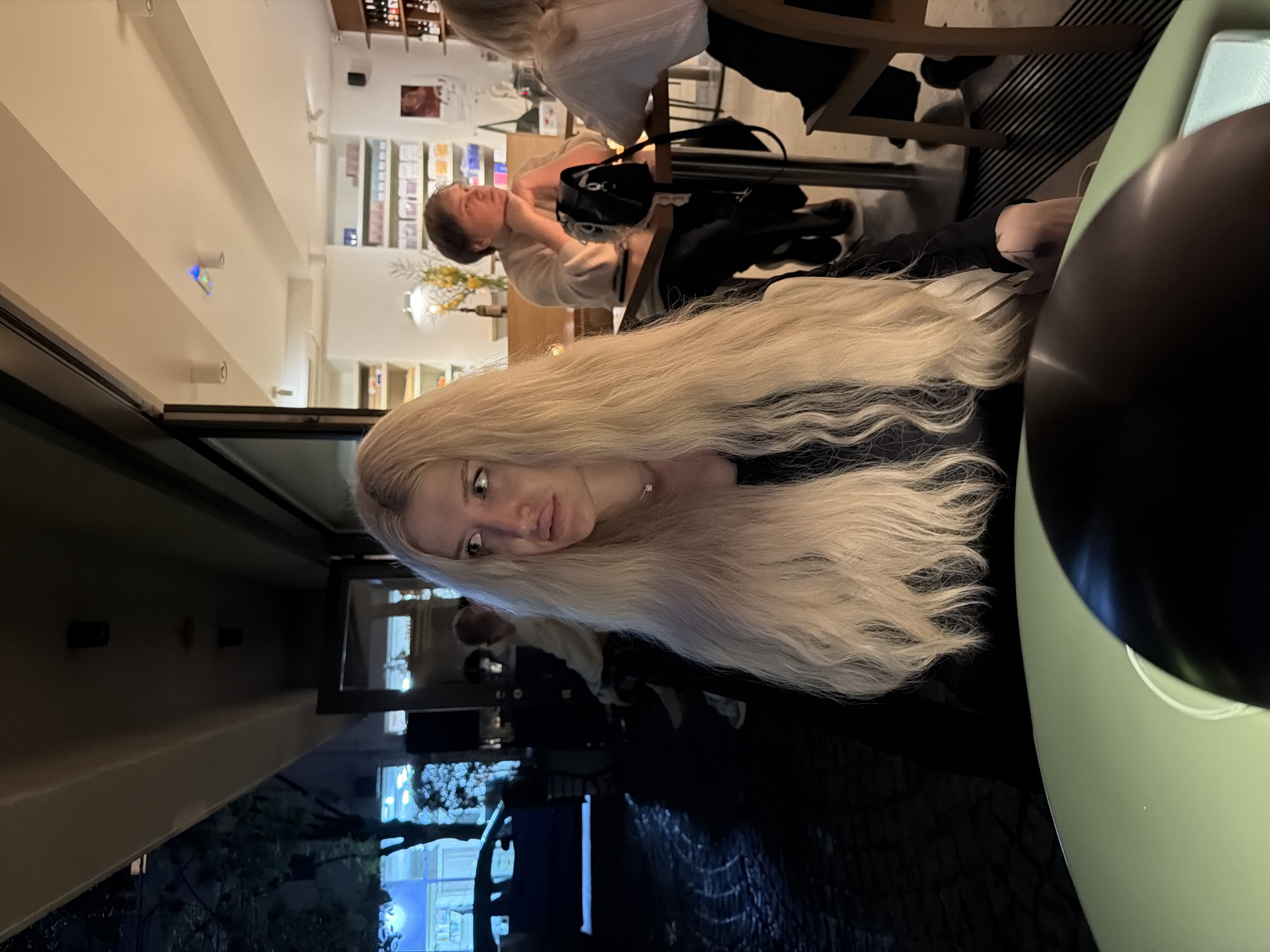 The image shows the interior of a restaurant or cafe in Belgrade, Serbia, at night. A blonde woman with long hair is looking at the camera. She is sitting at a table with a green surface. In the background, other patrons are visible. Through the window, the city lights of Belgrade are reflected.