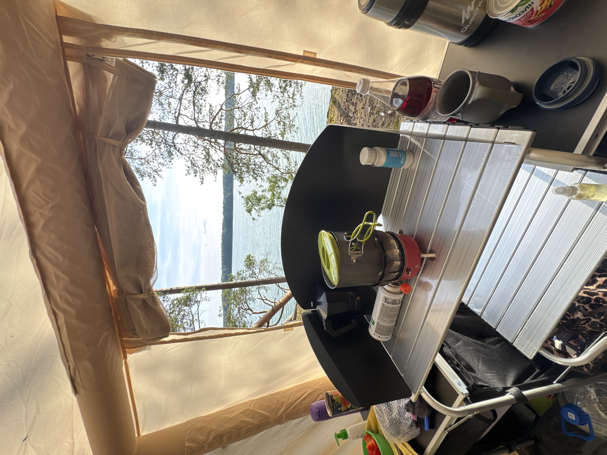 The image shows a campsite setup with cooking equipment and supplies laid out on a portable table in front of a tent. The scene is set by a body of water, likely a lake, bordered by trees, suggesting a natural outdoor environment in the vicinity of Priladozhskoe, Russia.
On the table, there is a portable stove with a pot on top, a moka pot, a fuel canister, a bottle of dark red liquid, a metal mug, and a small container. To the left, there is a green bowl with red chopped vegetables, a bottle of soap or sanitizer, and a rolled-up towel. A thermos is also visible on the right side of the table, along with a can of food. The table itself is made of a silver, slatted material. The tent fabric is visible at the top and sides of the frame, in a light beige color.
The background reveals a serene lake with calm water and a distant shoreline lined with trees. The sky is clear with some light clouds. The overall impression is one of outdoor recreation and self-sufficiency in a natural setting. No people are present in the image.