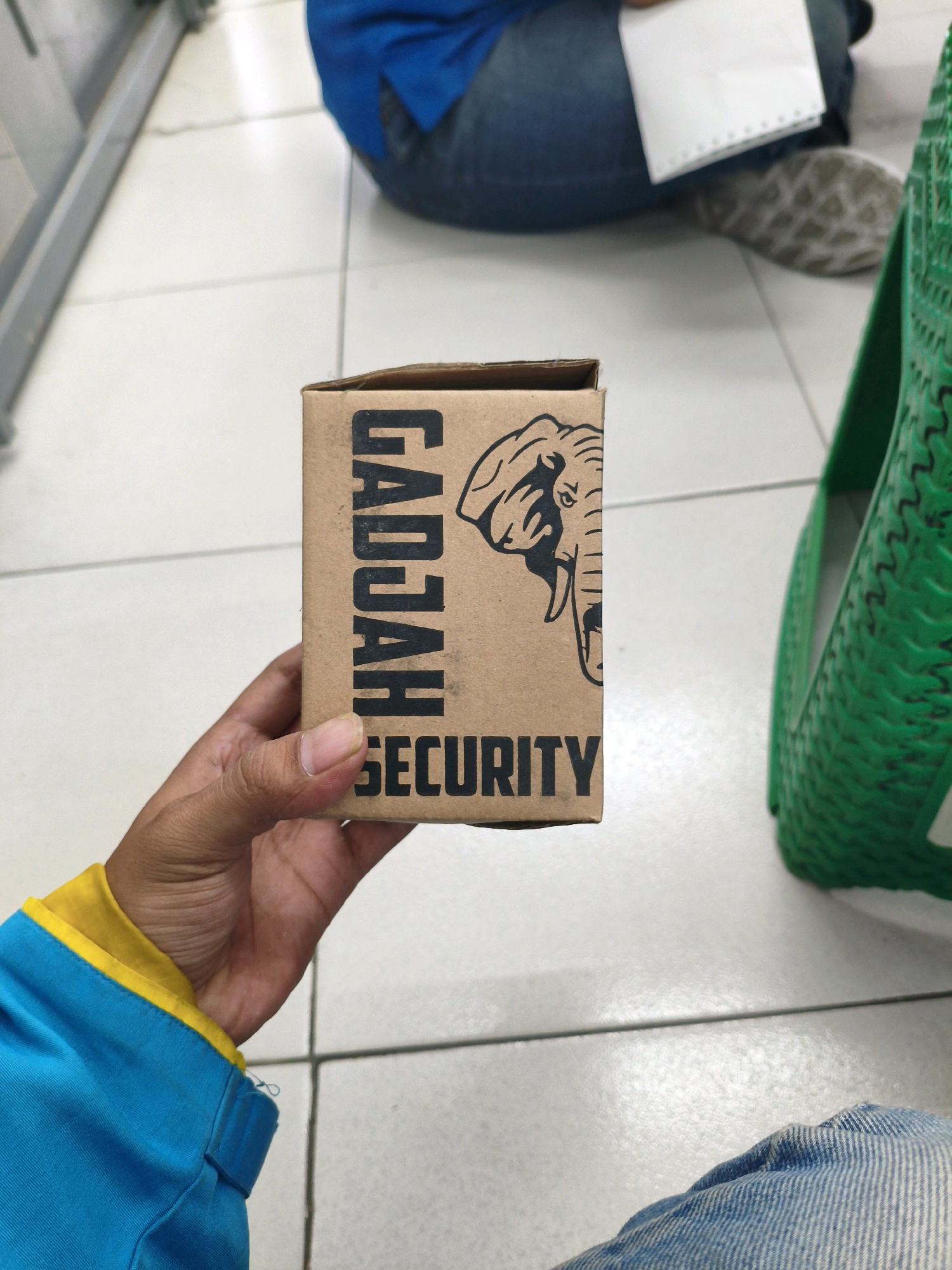 A person's hand, exhibiting brown skin and short fingernails, holds a small, rectangular cardboard box. The box is oriented vertically, displaying text and an illustration. The text reads "GADJAH" in large, bold, black, capital letters running down the left side. To the right of "GADJAH" is a black-and-white illustration of an elephant's head in profile, facing left, with prominent tusks and ears. Below the elephant's head, in smaller, black, capital letters, is the word "SECURITY." The box is a light brown, unvarnished cardboard.
The person is wearing a blue jacket with a yellow collar. The background shows a tiled floor, with visible grout lines. To the right of the box, a portion of a bright green, textured basket is visible. In the upper background, parts of two individuals are discernible: one wearing a blue shirt and jeans, seated on the floor, holding a white piece of paper with perforations along one edge. The other person, also in blue and jeans, is out of focus. The scene appears to be indoors, possibly in a public space like a store or waiting area. The image is sharp and well-lit. There is no specific information to indicate the location is Sumberejo Kulon, Indonesia.
