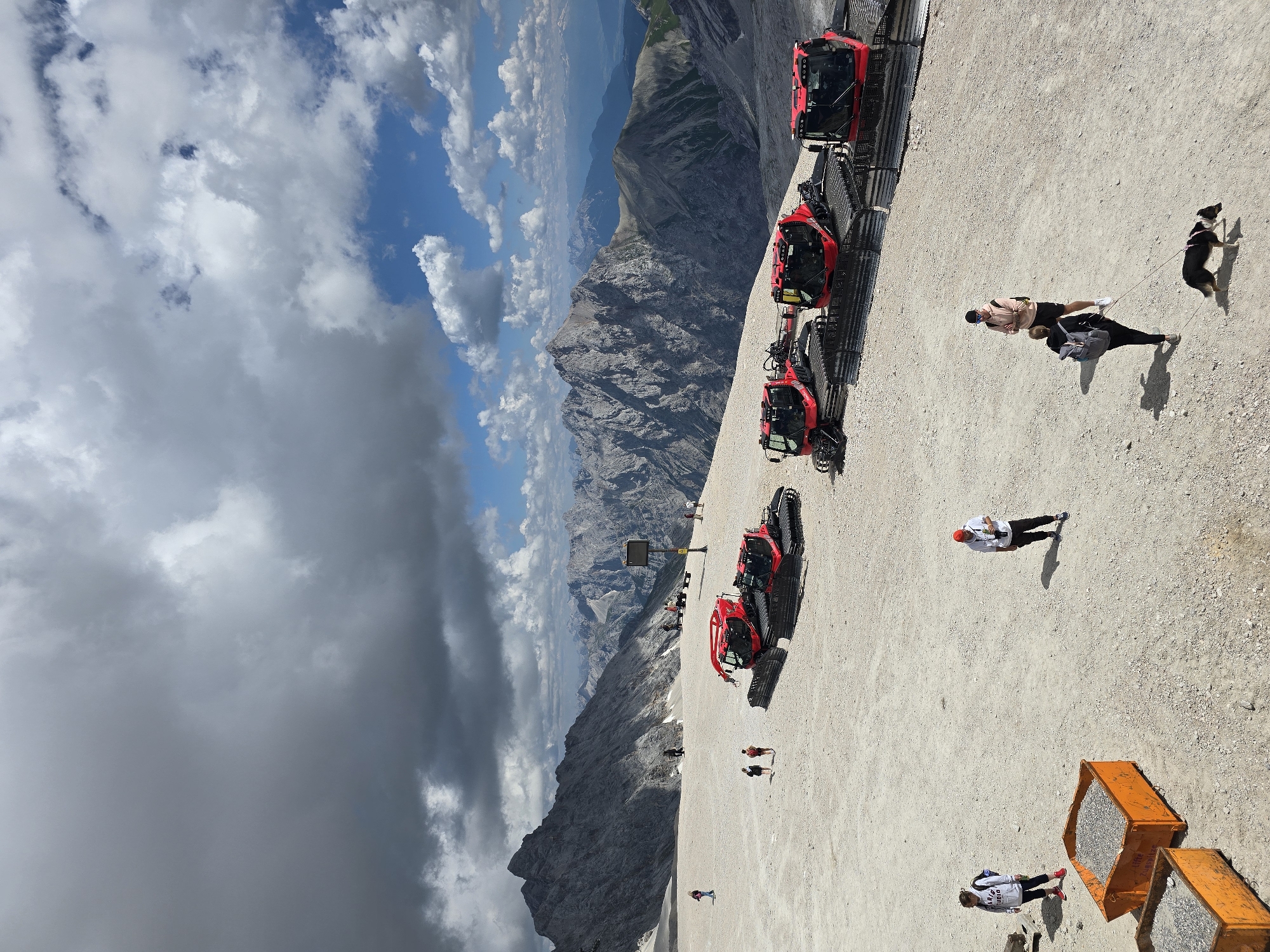 The image depicts a scenic mountain landscape with a clear sky and scattered clouds. In the foreground, there are several red snowcats parked on a gravelly surface. A few people are visible, some walking and one with a dog. In the mid-ground, more people are seen scattered across the area, with a dark, imposing mountain range in the background. The scene suggests a location in the Bavarian Alps, potentially near Garmisch-Partenkirchen, Germany, a region known for its mountainous terrain and winter sports facilities, although the presence of snowcats indicates a need for snow grooming even in non-winter months or at higher altitudes. The overall impression is one of a vast, rugged natural environment with signs of human activity.
