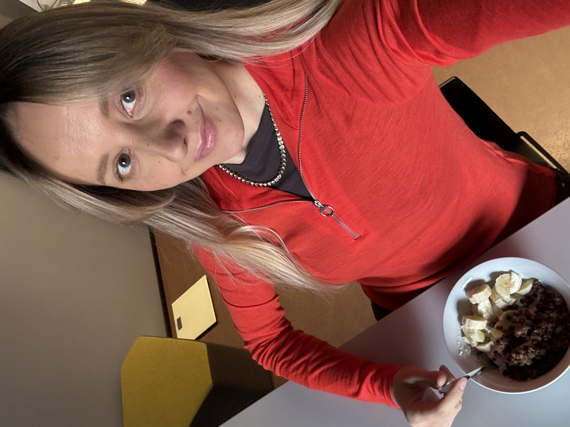 A high-angle selfie captures a woman looking directly at the camera with a slight, pleasant smile, taken indoors in Riga, Latvia. She has long, blonde and brown hair styled with bangs. She is wearing a bright red long-sleeved quarter-zip top over a black undershirt, accessorized with a silver chain necklace. Her right hand holds a spoon, positioned over a white bowl of food. The bowl contains a dark, granular dish, likely a type of cereal or oatmeal, topped with several slices of banana and a lighter, possibly creamy, sauce. The food and spoon rest on a light gray table. The background is a subtly lit indoor setting, showing muted-colored walls and soft hints of furniture, including a yellow geometric accent. The scene depicts a casual moment, possibly during a meal or snack. No text is visible in the image.