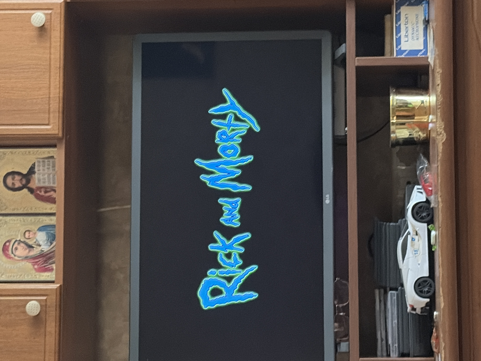 The image captures a television screen displaying the "Rick and Morty" logo, rendered in vibrant blue and green with a stylized, jagged font. The television itself is a flat-screen model with an LG logo visible at the bottom right corner. It is positioned on a wooden shelf unit. Above the television, on a higher shelf, are two framed religious icons. The icon on the left depicts a woman in a red headscarf holding a child, and the icon on the right shows a man with a beard. The shelf unit also contains various objects, including a white toy sports car with the number '18' on its side, a brass cylindrical container, and a blue box labeled "Liberton DYNAMIC MICROPHONES." The overall setting appears to be a home entertainment area.