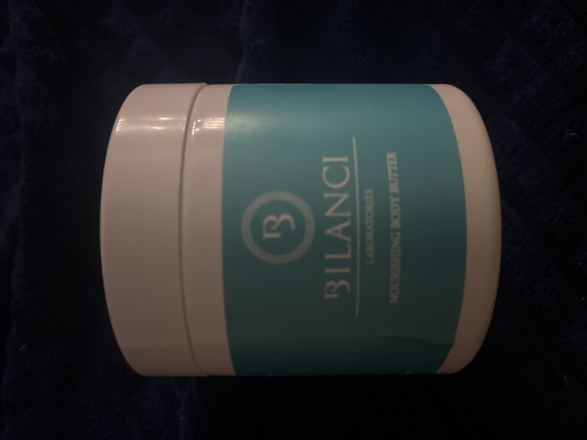 A white jar of "BILANCI LABORATORIES NOURISHING BODY BUTTER" is displayed on a dark blue textured surface. The jar has a teal label with white text and a circular logo containing the letters "B". The lighting suggests an indoor, possibly domestic, setting. There are no people or animals visible in the image. The location in the city of Volsk, Russia, is not discernible from the visual information provided in the image.