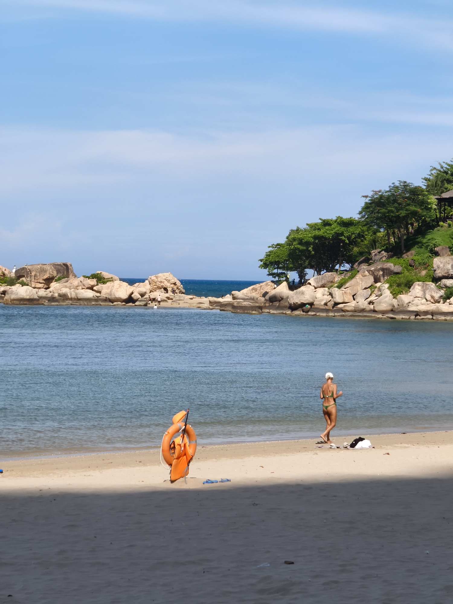 The image captures a section of a sandy beach in Nha Trang, Vietnam, with calm, blue ocean water. In the foreground, a bright orange life preserver is propped up on the sand. Further back, a woman wearing a green bikini and a white cap is walking away from the camera towards the water. A towel and clothing are laid out on the sand near her. The background features a rocky coastline with lush green trees and a small structure on the hill, possibly a gazebo or a hut. The sky is clear blue with faint clouds.