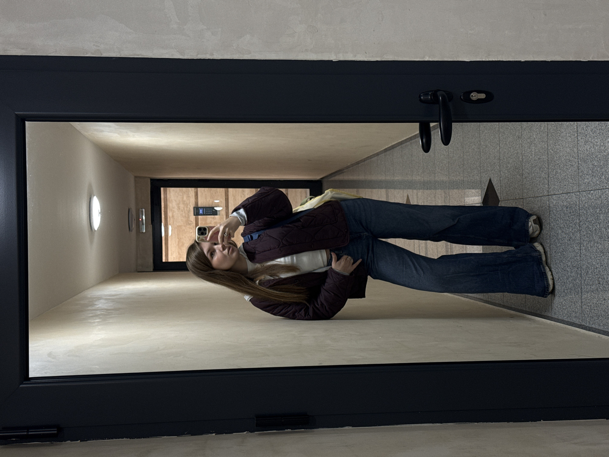 A young woman is reflected in a large mirror framed by a black door frame. The reflection shows her standing in a hallway. The hallway has beige walls with evenly spaced, round light fixtures. The floor is a gray, tiled surface. She is wearing blue jeans, a white top, and a dark purple jacket. She holds a smartphone in her right hand, taking a selfie. The reflection extends deep into the hallway, showing another doorway at the end. The scene appears to be indoors, possibly in an apartment building or office space. The location, Vozdovac Urban Municipality, Serbia, is impossible to confirm visually.