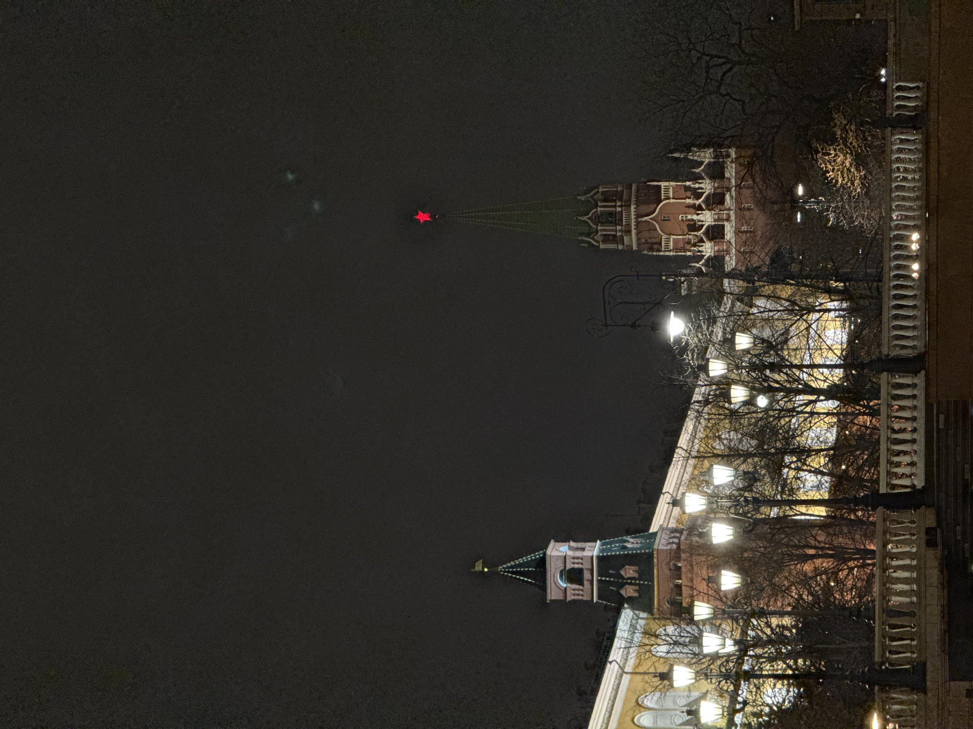 At night, the image shows the Moscow Kremlin. Two towers are visible, one topped with a red star. A row of streetlights illuminates trees in front of the buildings. The sky is dark.