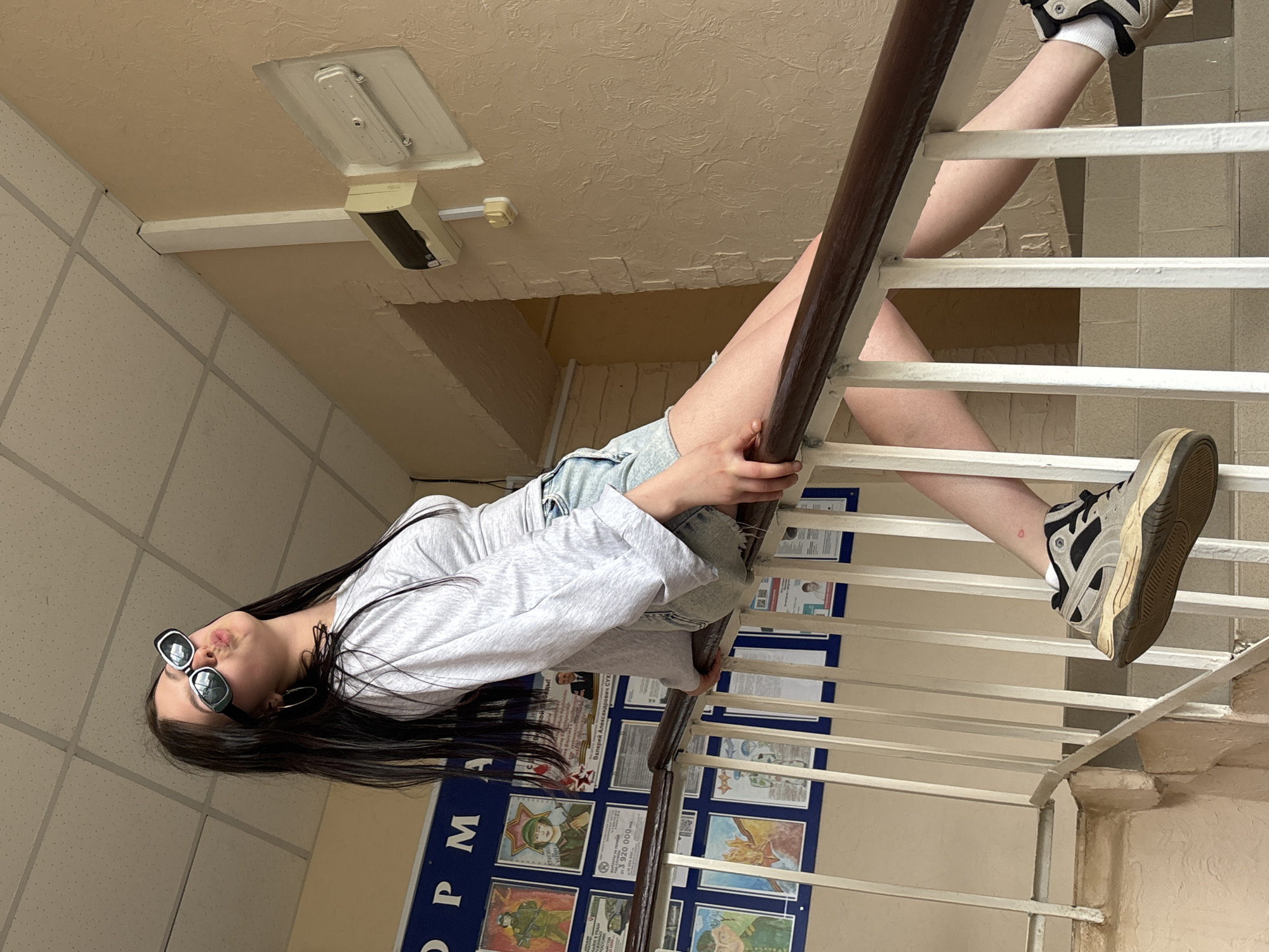 A person, possibly female, is sitting on a stair railing in an interior space. The space appears to be in Фоки, Russia, evidenced by the presence of Cyrillic writing on a board behind the railing. The person is wearing a gray sweatshirt, jean shorts, and sneakers with white socks. They are also wearing sunglasses and have long, dark hair. The walls are painted a neutral color. The stair railing is white with a brown handrail. The person is holding onto the handrail and has their legs extended. A noticeboard is behind the railing and displays information, including images and text in Cyrillic. The ceiling is a mix of tile and plaster, and contains lighting fixtures.