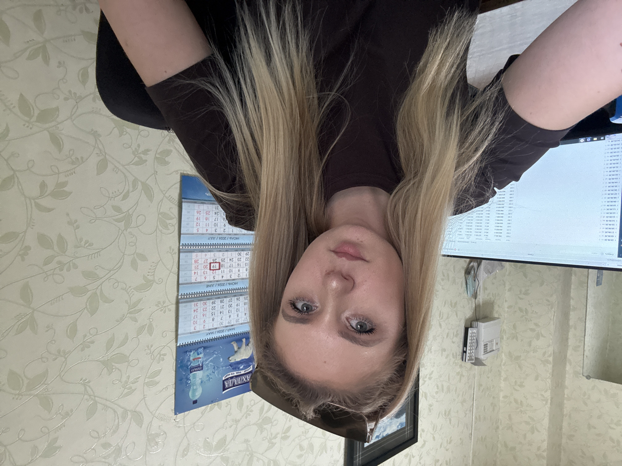 A young woman with blonde hair and blue eyes is looking at the camera. She is wearing a dark brown t-shirt. She has sunglasses on top of her head. In the background, there is a calendar with the months of May, June, and July 2026. There is also a computer monitor displaying data, and a wall with floral wallpaper. The image appears to be taken indoors, possibly in an office or home environment.