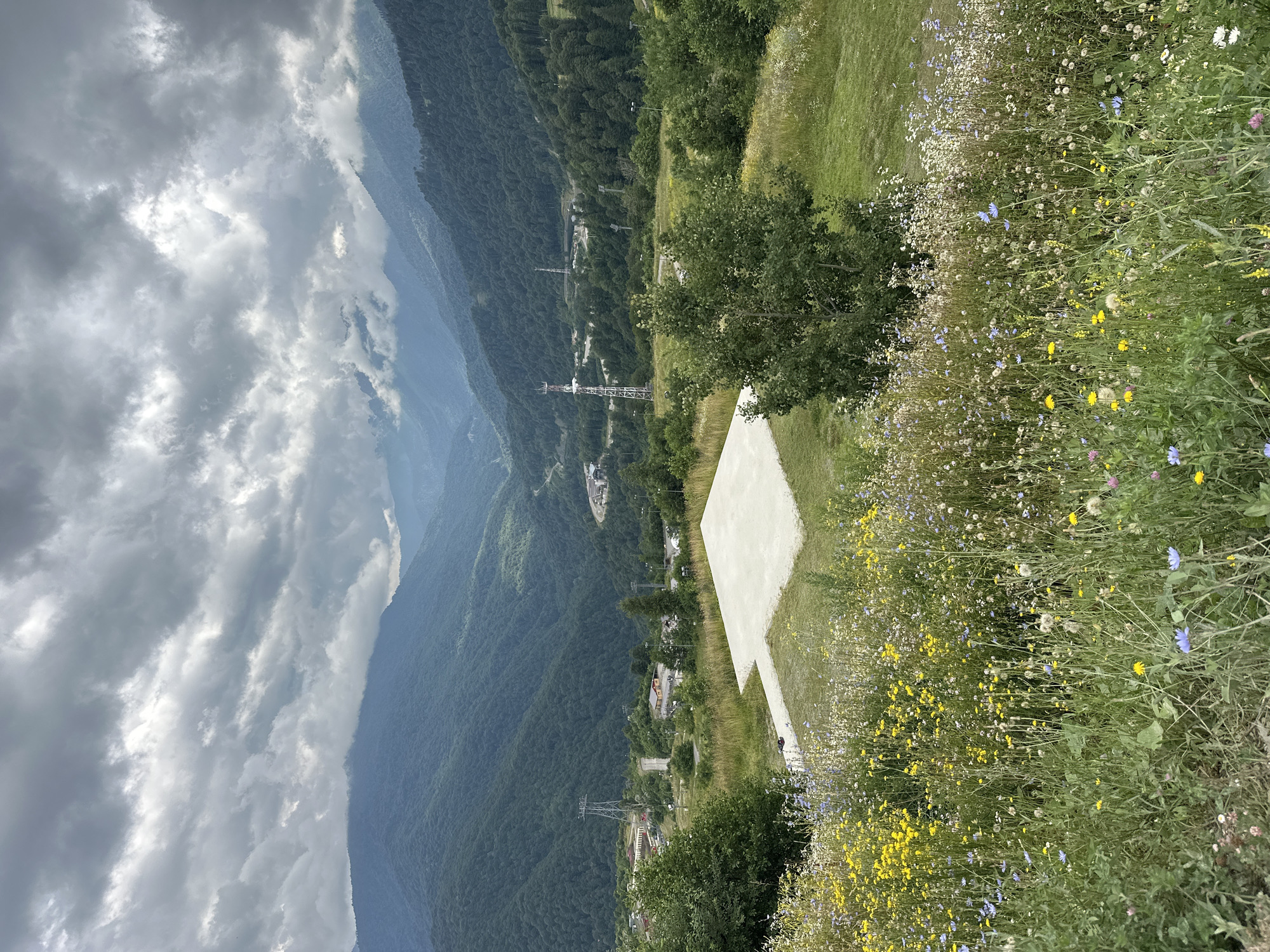 The image shows a scenic mountain landscape with a cloudy sky above. In the foreground, a patch of wildflowers with yellow, blue, and white blooms covers a grassy slope. A large, light-colored concrete helipad or landing pad is visible on the grass in the mid-ground. Beyond the helipad, a valley stretches out, dotted with buildings, roads, and utility towers. The valley is surrounded by lush, green, tree-covered mountains that recede into the distance. The sky is filled with dramatic, layered clouds, with some patches of sunlight breaking through, illuminating parts of the mountains. This scene is consistent with the mountain resort area of Estosadok, Russia, known for its natural beauty and recreational facilities. There are no people or animals visible in the image.