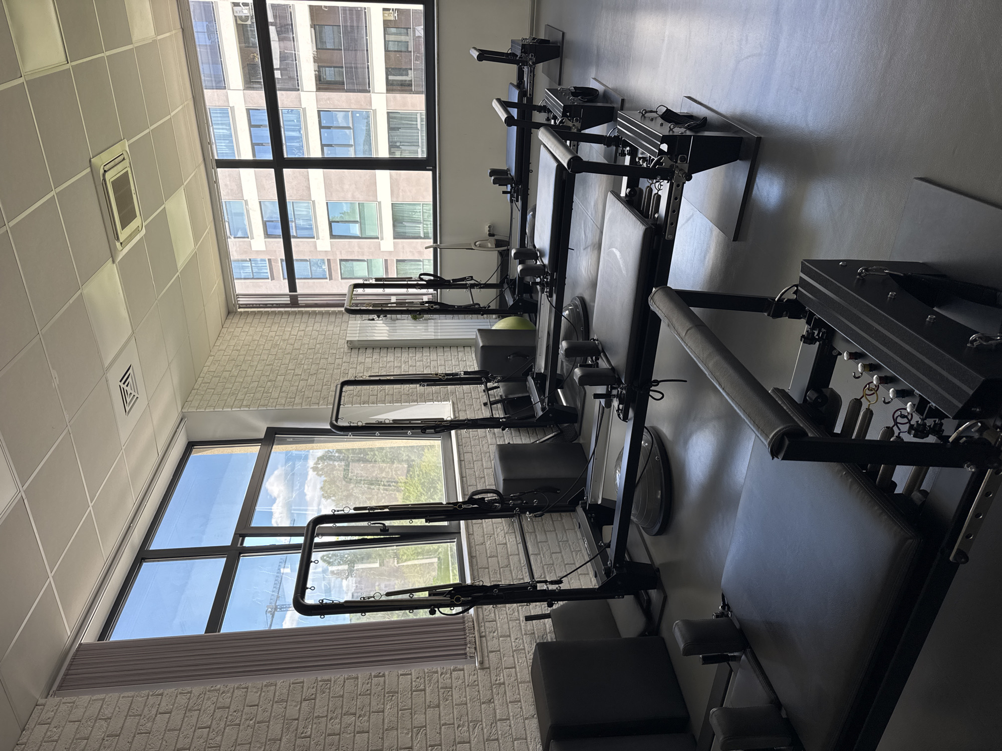 The image shows a pilates studio in Sofia, Bulgaria. Several pilates reformer machines are arranged in a row, oriented towards large windows that offer views of neighboring buildings and the cityscape. The studio walls are a light brick, and the flooring appears to be a smooth, neutral-colored material. The ceiling is a grid pattern. There are no people visible in the frame.
