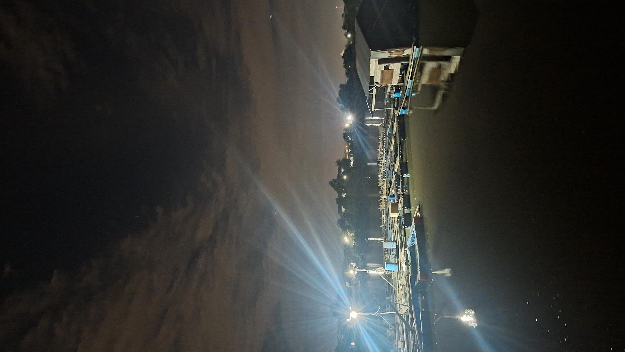 The image depicts a nighttime scene on a body of water, likely a river or lake, in Lhokseumawe, Indonesia. The foreground is dominated by dark, reflective water, with small white specks scattered across its surface, possibly reflections or debris. In the mid-ground, a series of floating structures are visible, resembling fish farms or aquaculture platforms. These structures consist of interconnected rafts with blue barrels and wooden planks. A spotlight on a tripod illuminates a section of these rafts, casting bright beams of light that spread across the water and into the dark sky, creating a starburst effect. In the background, a shoreline with vegetation and buildings is dimly visible, with additional lights indicating a populated area. A small, dilapidated wooden structure on a raft is present on the right side of the image, showing signs of wear and tear. The sky above is dark with scattered clouds, some of which are illuminated by the artificial lights. The overall impression is one of a functional, working waterfront at night, possibly a fishing or aquaculture operation.
