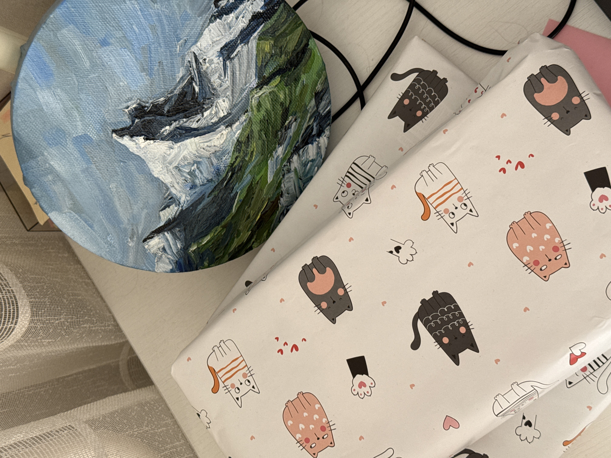 A high-angle shot captures an indoor scene featuring a round canvas painting, a stack of gifts wrapped in paper with a cat pattern, and a portion of a window.
The painting depicts a landscape, likely mountains, with snow-capped peaks painted in white and gray, set against a blue sky. Green vegetation covers parts of the mountain slopes.
Two wrapped gifts sit adjacent to the painting. The wrapping paper is white with a repeated cartoon cat design in various poses and colors (black, orange, pink). Small hearts are scattered around the cats.
In the lower left corner, a partially visible beige curtain with vertical pleats hangs near the painting and gifts. A black cord or cable runs across the top of the gifts.
The scene is well-lit, suggesting natural light through the window. The setting appears to be a room within a home or shop. There's no indication of the specific location in Krasnoyarsk, Russia.