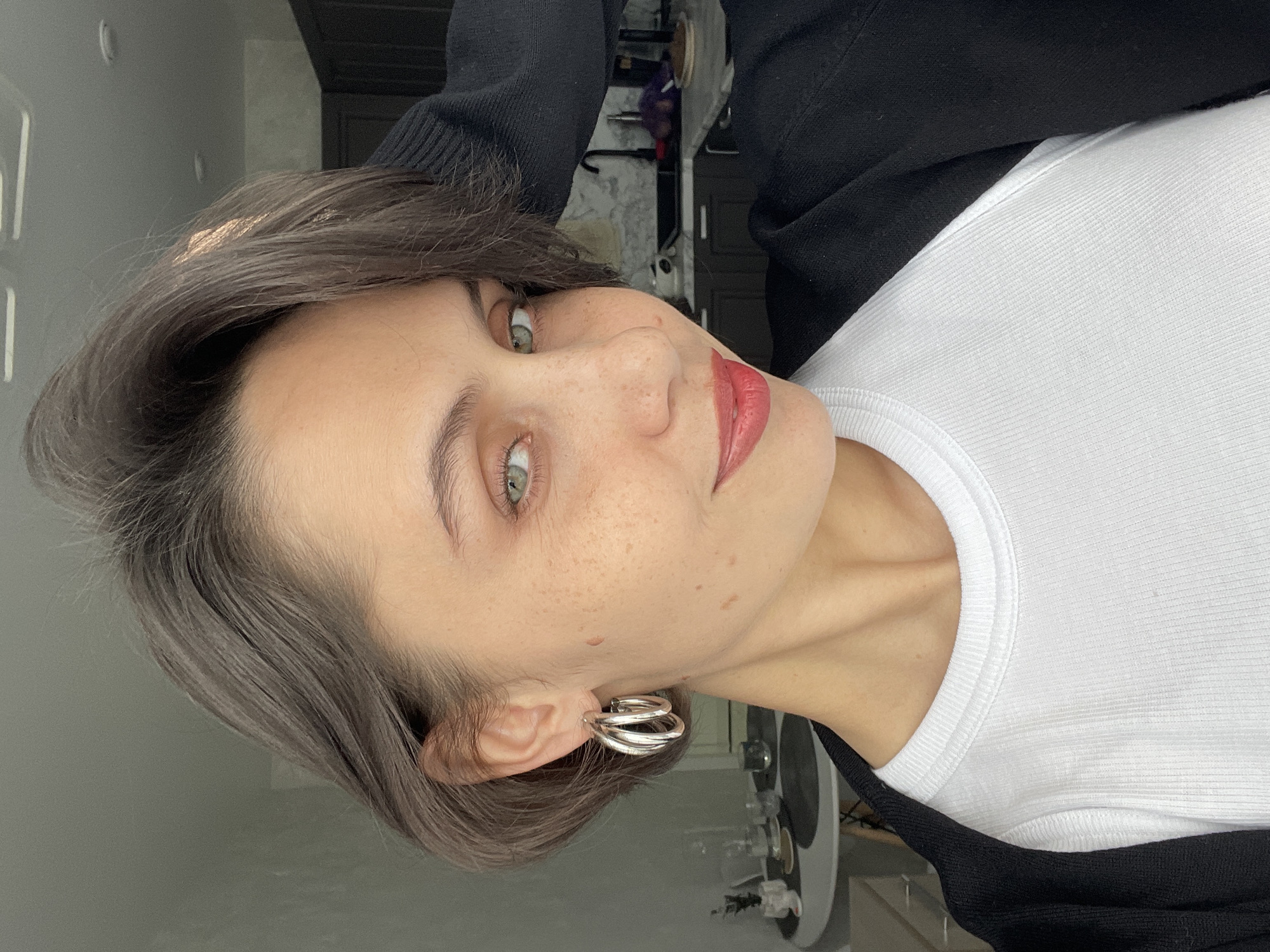 A woman is photographed in a close-up, tilted portrait shot. She has short, grey-brown hair, fair skin with freckles, and light green eyes. She is wearing silver hoop earrings and has red lipstick. The woman is wearing a white ribbed tank top layered under a black sweater. The background is a blur of light-colored walls and dark furniture. There is no indication of the location being Izhevsk, Russia.