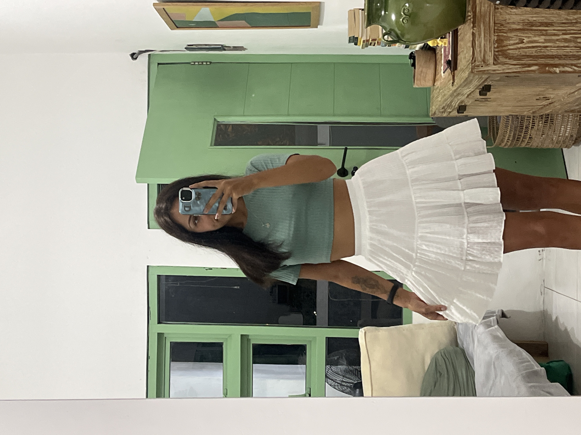 A young woman, likely in her 20s, is captured in a mirror selfie, standing indoors. The image is oriented vertically (portrait mode). She has long, dark hair and is holding a smartphone with a light blue patterned case, which reflects part of her face as she takes the picture. She is wearing a light mint green, short-sleeved crop top that exposes her midriff, paired with a white, tiered, ruffled mini-skirt. A black fitness tracker or watch is visible on her right wrist, and a tattoo is present on her right forearm.
Behind the woman, the room features a prominent sage green-painted wooden structure, possibly built-in cabinetry or a window frame, with dark windowpanes that do not reveal the outside environment clearly. Above this green structure, a cream-colored wall displays a framed abstract artwork with geometric shapes in earthy tones of yellow, green, and brown. To the right, a rustic wooden cabinet or side table is visible, topped with a large, olive-green ceramic pot or vase. Below the cabinet, a woven basket can be seen. In the lower right corner, part of a light-colored armchair or sofa with a cushion is visible, alongside some draped fabric in green and light grey/white, and a partially visible black fan. The overall setting suggests a cozy indoor space, possibly in Kuta Selatan, Indonesia, though no specific geographical markers are present within the image itself. The lighting appears to be ambient indoor light.