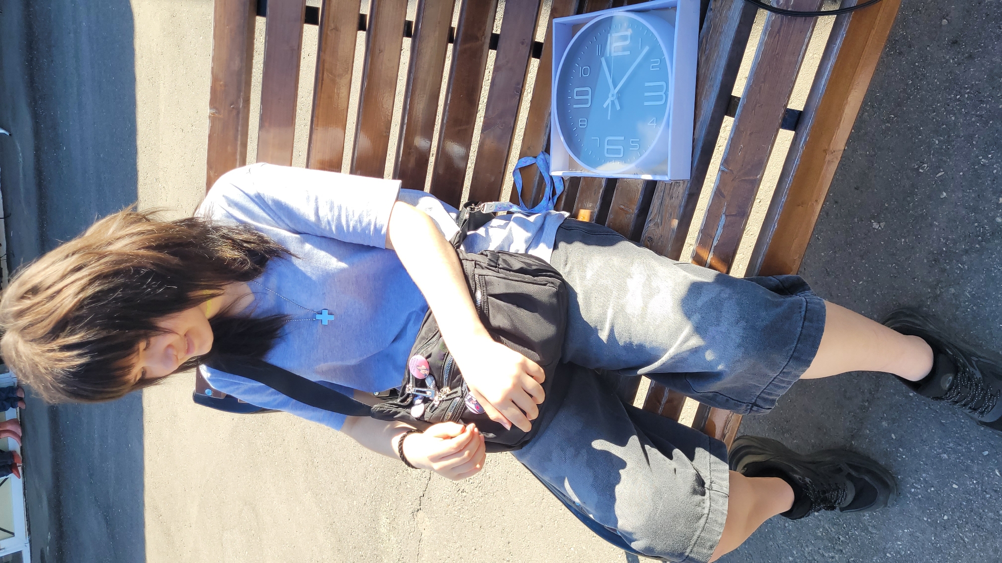A young woman is reclining on a wooden bench, positioned horizontally in the frame. She is wearing a light blue t-shirt, dark grey shorts, and black shoes. A black bag is slung across her chest, and a silver cross necklace is visible around her neck. Her dark hair obscures part of her face as she looks down. Beside her, on the bench, is a boxed white-framed clock. The bench is set against a grey paved surface. The location is an outdoor area in an urban environment, with the city location remaining Unknown.
