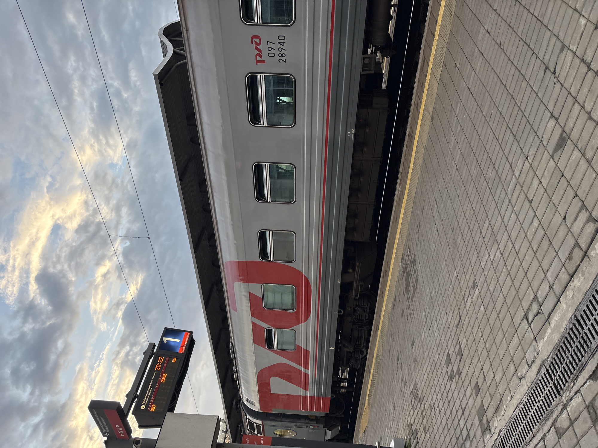 An outdoor, eye-level shot captures a train at a station under a cloudy sky. The train car is predominantly grey with large, red Cyrillic lettering on its side. A digital display board shows train information, including times and platform numbers, with the number "1" prominently displayed in a blue square. The platform itself is paved with grey square tiles and features a yellow line marking the edge. A metal grate is visible along the left side of the platform. The sky is filled with textured clouds, illuminated by sunlight breaking through. The scene appears to be in Khabarovsk, Russia, indicated by the train's markings and the Cyrillic text on the display board.