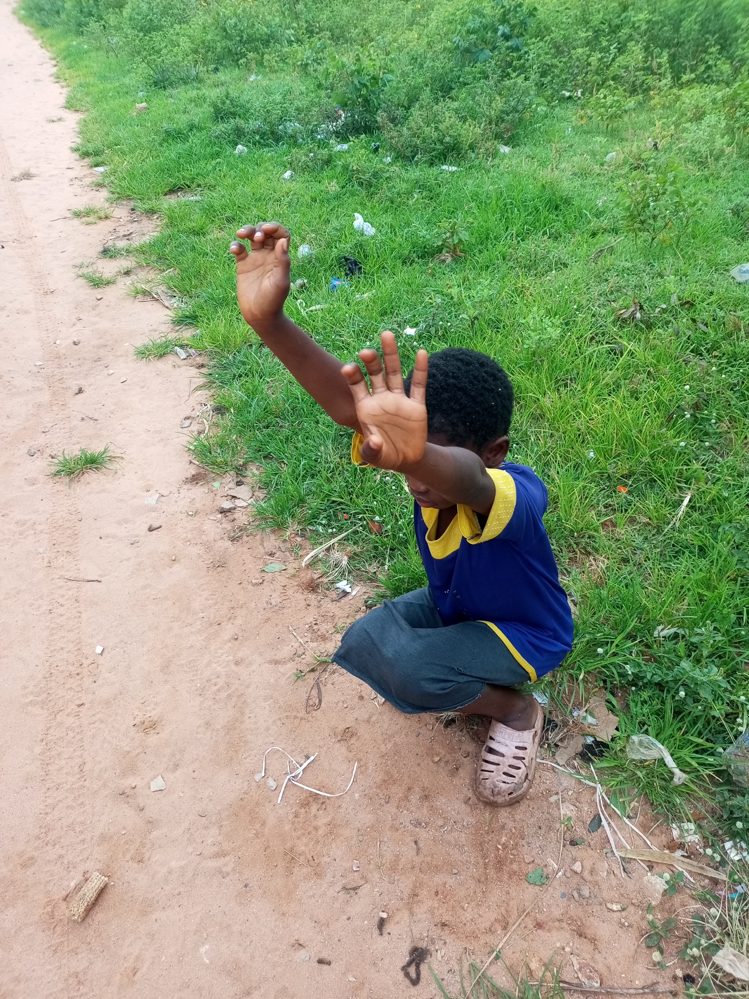 A young Black child, wearing a blue t-shirt with yellow trim and dark pants, crouches on a dirt path adjacent to a grassy embankment. The child's hands are raised in front of their face, palms facing outward, in a gesture that could indicate surprise, fear, or refusal. The child is wearing light-colored, open-toed sandals. The environment appears to be outdoors, with a dirt path on the left and lush green grass, interspersed with some scattered debris, on the right. The scene is set in Tempure, Nigeria.