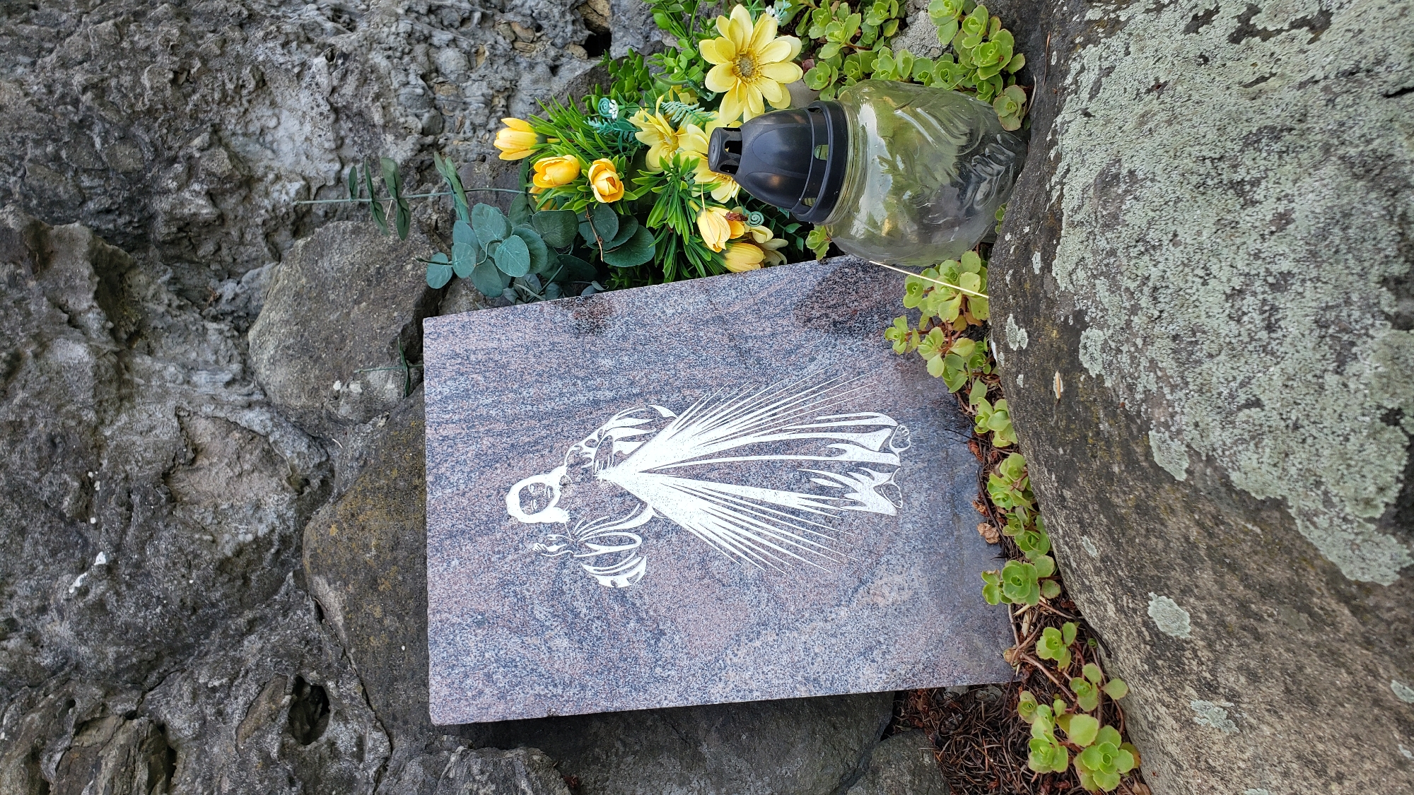 The image shows a memorial plaque with a depiction of Jesus Christ, standing upright against a rough, stone wall. The plaque is made of polished granite with a marbleized pattern and features a white outline of the figure. To the right of the plaque, a cluster of yellow artificial flowers and a clear glass grave candle with a black lid are placed on rocks. Small green succulents are growing around the base of the plaque and the grave candle. The background consists of large, weathered stones and some greenery. The scene is situated outdoors, likely in a cemetery. The location is specified as being in the city of Prešov, Slovakia.