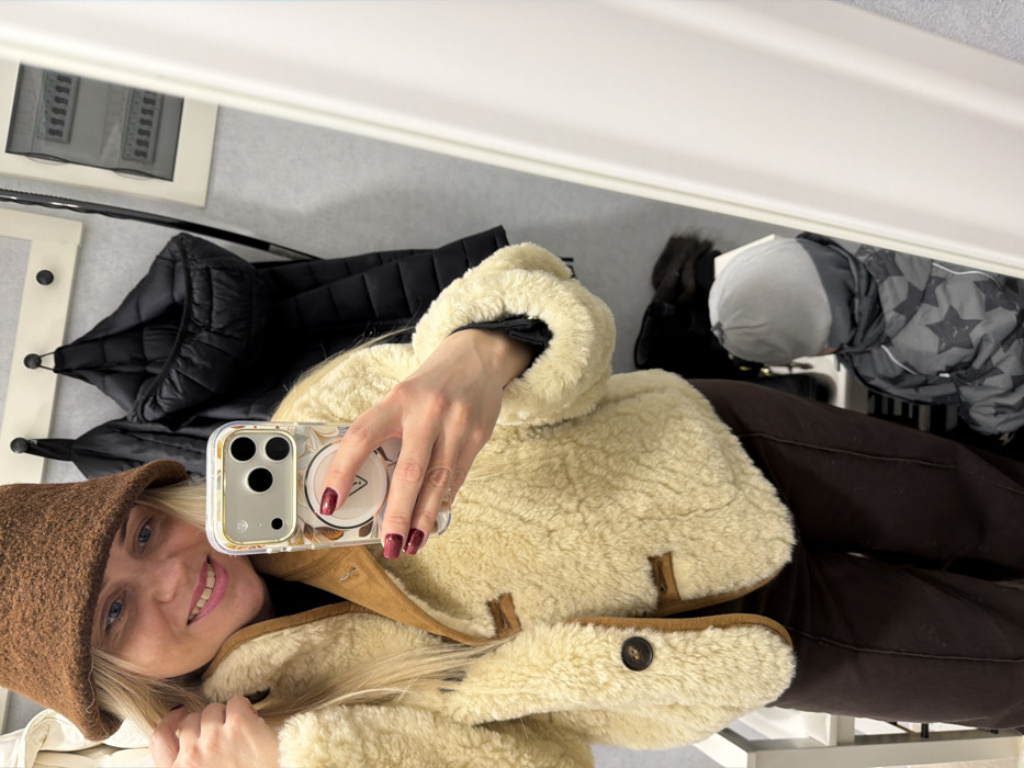 This image depicts an indoor scene within what appears to be a changing room or retail environment in Votkinsk, Russia. The central figure is an adult female with blonde hair, wearing a brown felt bucket hat, a cream-colored shearling-style jacket with brown trim and dark brown toggle buttons, and dark brown trousers. She is smiling directly at the camera, holding a smartphone with a patterned case and dark red manicured nails in her left hand, indicating a mirror selfie. Her right hand is touching her hair near her right temple. In the background, a light-colored wall displays a white coat rack with three black hooks, from which a black puffer jacket is suspended. A framed object, possibly an electrical panel or ventilation system with horizontal slats, is visible on the wall to the right. Reflected in the lower right portion of the mirror is a small child, wearing a grey beanie and a grey winter jacket with a black star pattern, seated on a low surface and partially obscured by the woman's reflection.