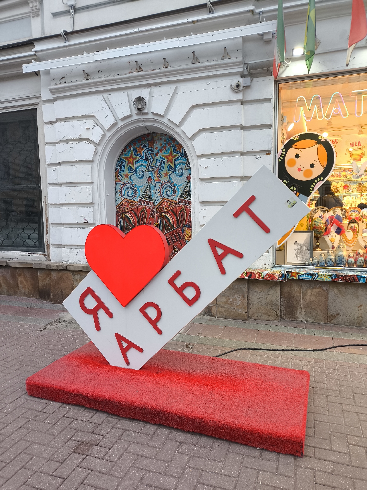 A street-level shot shows a large, red heart leaning against a white sign with Cyrillic red letters. The sign stands on a rectangular red platform. The sign reads "Я Арбат," likely a reference to Arbat Street in Moscow, Russia. The background features a building with a decorative mosaic panel above a window on the left. To the right is a shop window displaying a Matryoshka doll. Flags of different colors hang from above the window. The street is paved with brick. The scene takes place during the day.