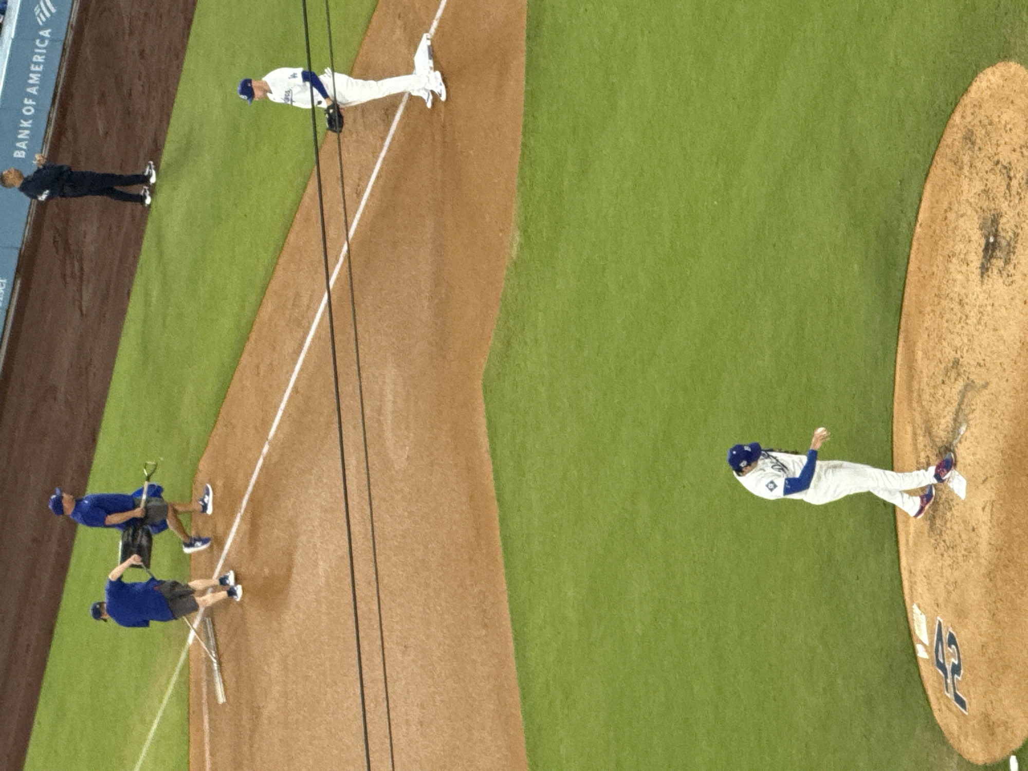An aerial view of a baseball field in Los Angeles, California shows a pitcher on the mound, about to throw a baseball. There are two other players on the field, standing at the first base line. Another man stands on the green grass behind the first base line.