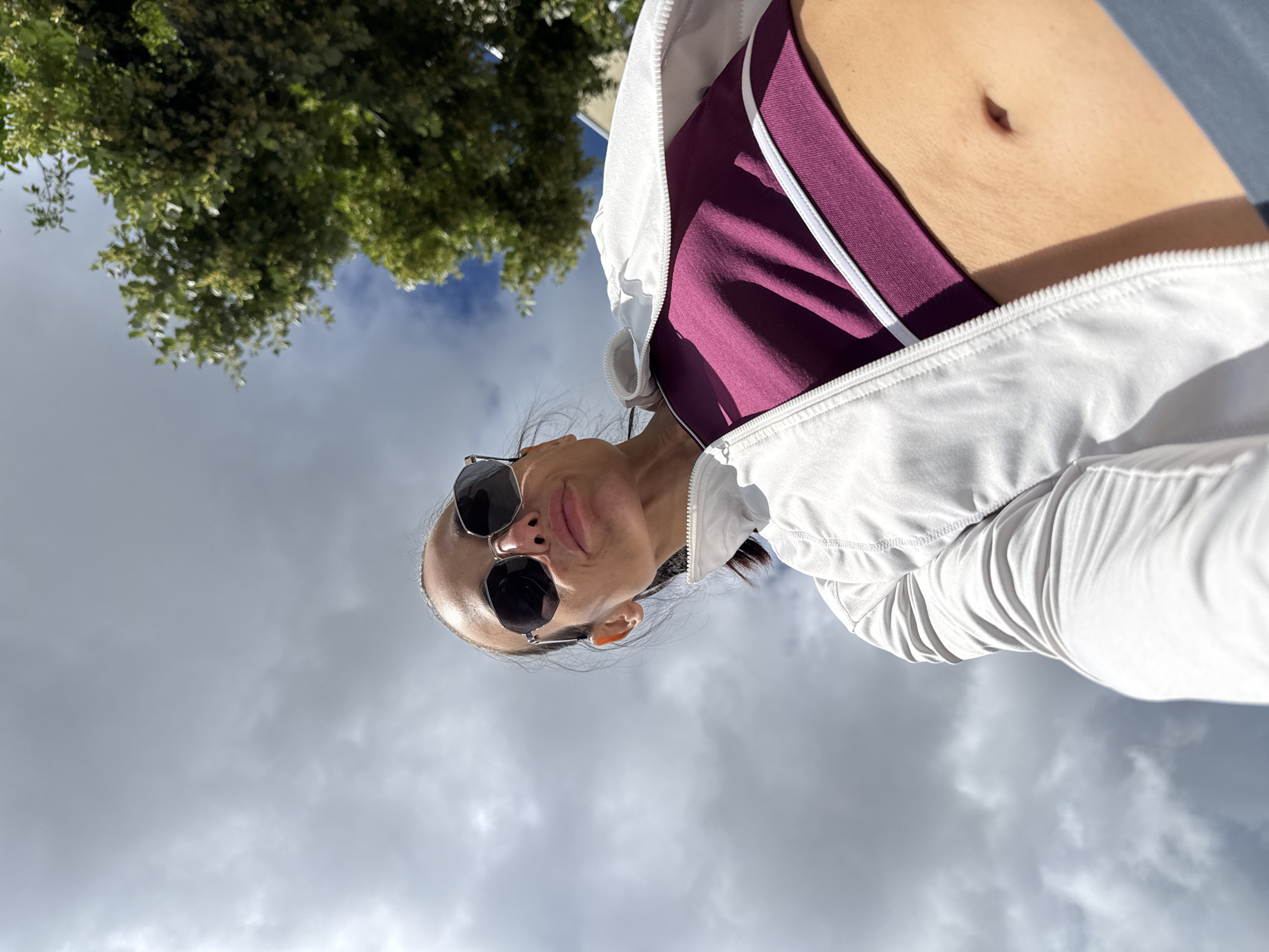 A low-angle, close-up, full-body shot taken outdoors in Pleven, Bulgaria, shows a woman from the chest up, against a background of cloudy sky and a tree. She is wearing a white zipped jacket over a burgundy sports bra and sunglasses. The woman is facing the camera with her head tilted upwards and slightly to the left. Her hair is pulled back into a ponytail, and she is wearing dark-rimmed, octagonal sunglasses. The image appears to be a selfie, taken with the camera held at a low angle, looking up at the woman. The sky is partially obscured by a green tree on the right side of the frame.