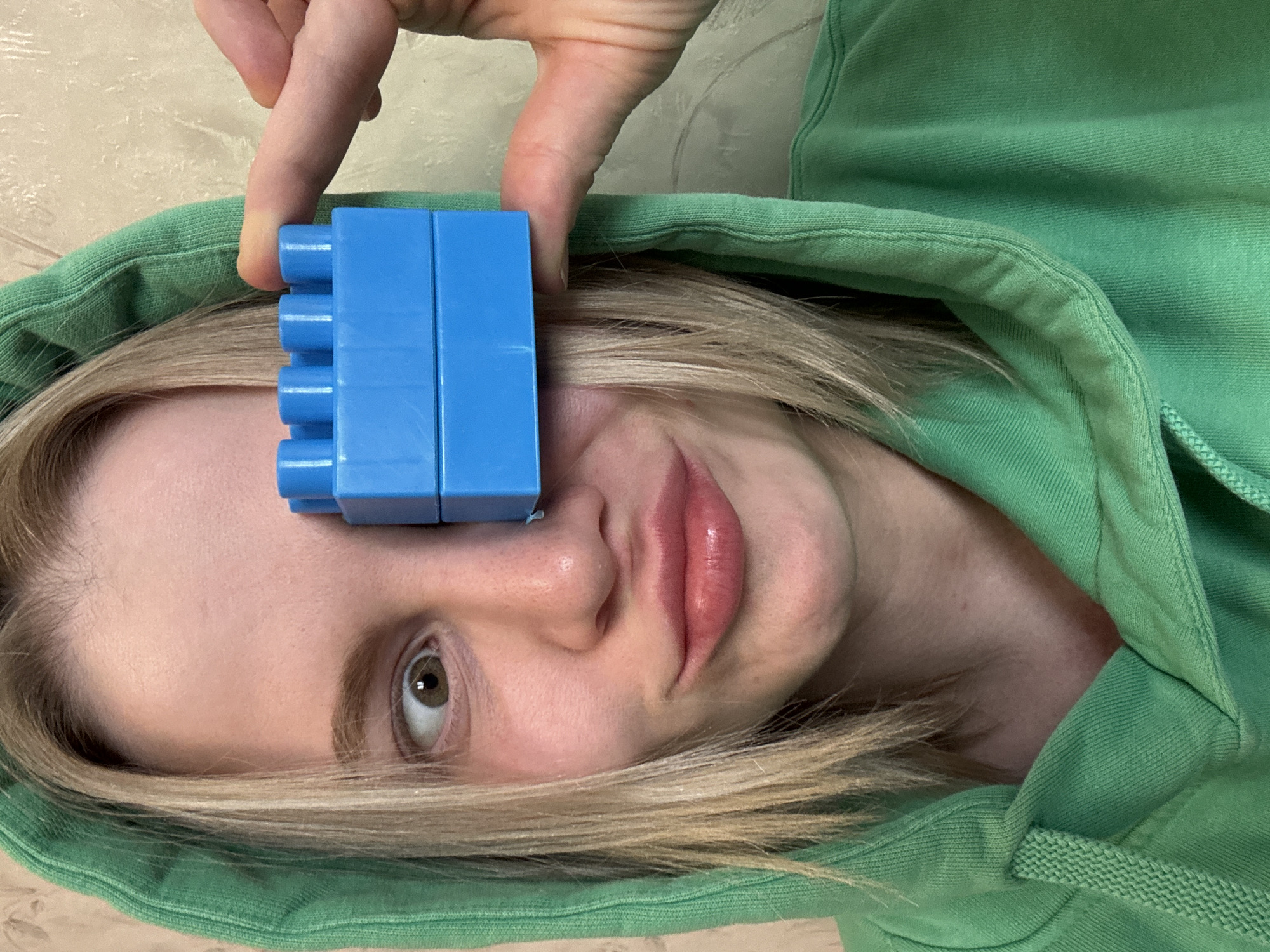A woman with blond hair and wearing a green hoodie is lying on her back. She has a blue building block over one eye, held in place by her right index finger. The background appears to be a textured surface. There is no visible evidence indicating a location in the city of Saint Petersburg, Russia.