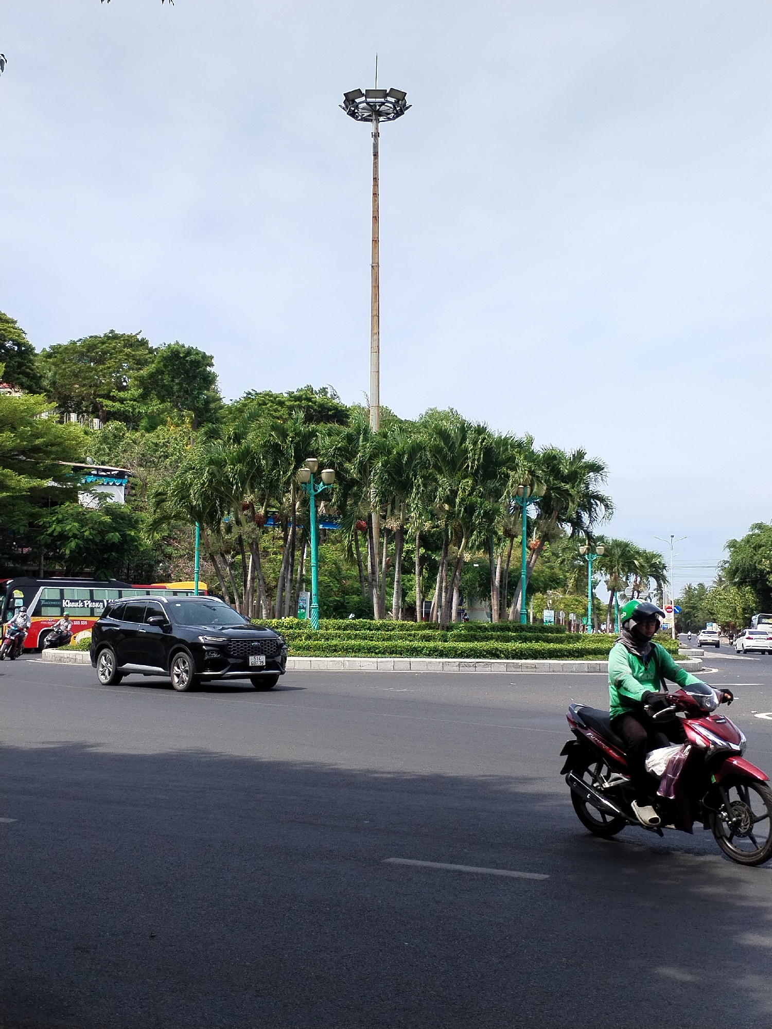 The image depicts a street scene in Bac Nha Trang Ward, Vietnam, on a clear day. In the foreground, a person wearing a green long-sleeved shirt and a helmet rides a red motorcycle. To the left, a black SUV with a license plate reading "51C 697.75" is traveling on the road. Behind the SUV, a red and black bus with the text "Khanh Phong" is visible. The background features a lush green area with numerous palm trees and other foliage, a large street lamp, and more trees on a hill. The sky is bright and cloudless.
