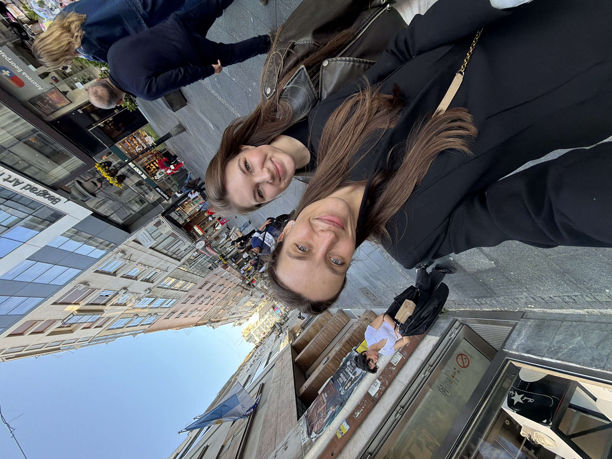 Two women are taking a selfie in a European city. They are wearing black outfits, one with a leather jacket and the other with a black blazer. The background shows a bustling street with shops, cafes, and buildings. Signs in the background mention "Beograd Art Hotel" and "Апотека Београд," suggesting the location is in Belgrade, Serbia. People are walking in the background, and the sky is visible above the buildings. An Argentinian flag is visible on the side of the building. A shoe store can be seen along the bottom right side of the photo.