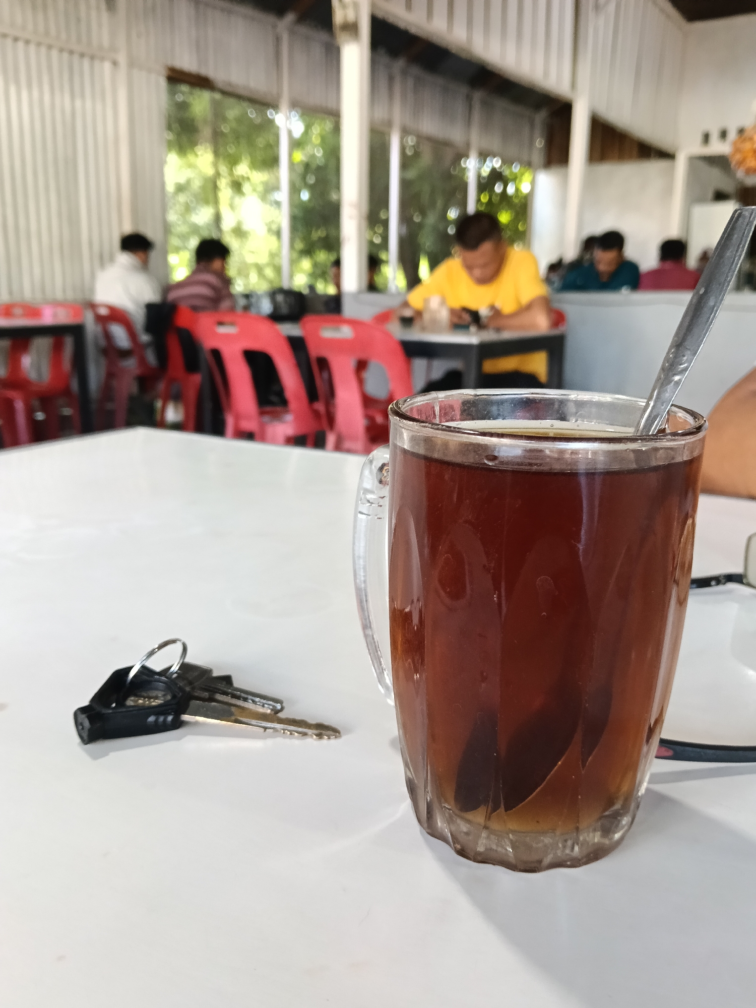 A glass mug of dark tea with a spoon is placed on a white table in what appears to be a casual eatery. In the foreground, a set of keys with a black fob and a silver ring rests to the left of the mug. In the background, several people are seated at tables, some looking at their phones. The environment suggests an indoor setting with a view of greenery through a window. The overall scene depicts a moment of pause and relaxation. This type of setting is common in many eateries and coffee shops found in Banda Aceh, Indonesia, where individuals often gather for refreshments and social interaction.