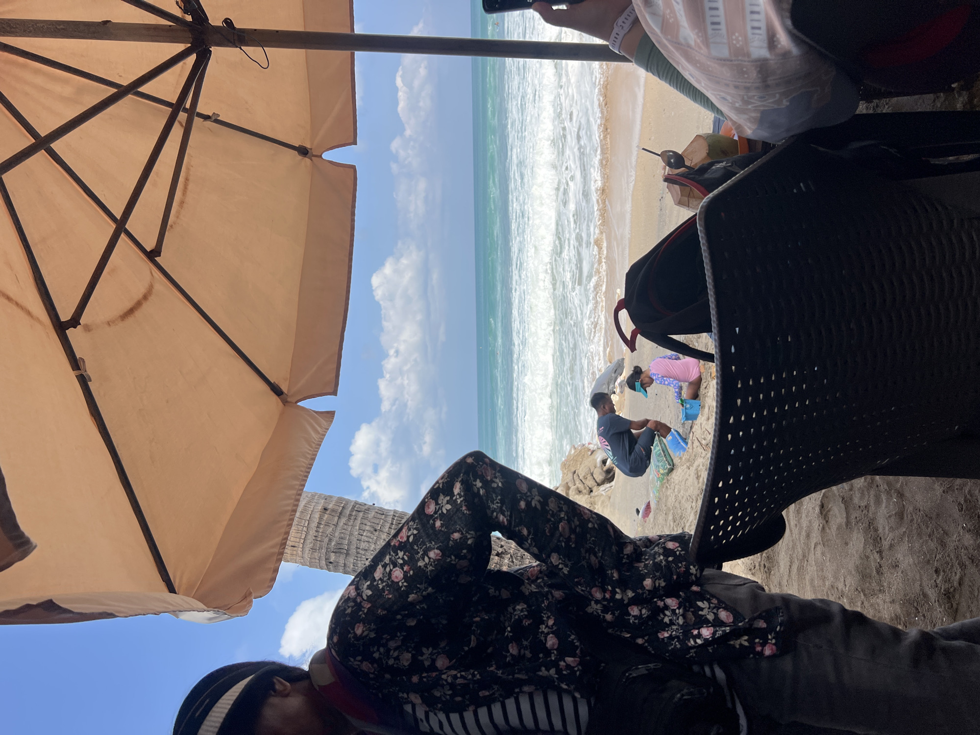 An outdoor scene on a sandy beach in Kuta, Indonesia features a group of people under a large, light-tan beach umbrella. In the foreground, a person wearing a dark floral patterned jacket and a striped shirt is partially visible, standing to the left. A black woven chair is in the center, and a table holds a coconut drink with a straw and a spoon, along with a backpack. In the background, two individuals are on the sand near the water's edge; one, a person wearing a dark t-shirt, appears to be tending to something on the ground, while a child in a pink long-sleeved rash guard and a blue visor is playing. The ocean is visible with moderate waves breaking on the shore under a bright blue sky with scattered white clouds. A palm tree trunk is also present behind the person in the floral jacket.
