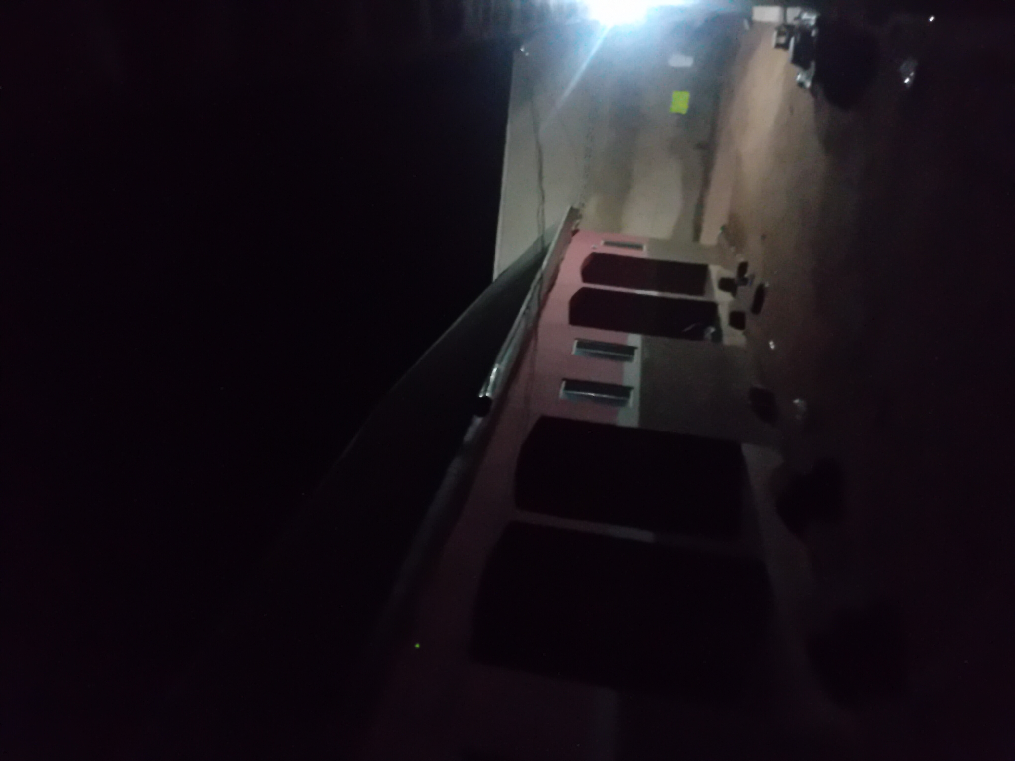 The image depicts an exterior view of a building at night in Jauro Yinu, Nigeria. The building appears to be a single-story structure with a pink and grey facade. It has a dark-colored roof that slopes downwards. Several dark, rectangular openings, likely doors or large windows, are visible on the front of the building. A bright light source is visible on the right side of the image, casting a glare and illuminating the surrounding area. The ground in front of the building is a mix of dirt and debris. Several dark, amorphous shapes, possibly objects or shadows, are scattered on the ground. The sky is dark, indicating nighttime. The scene appears to be a residential or commercial area within the city.