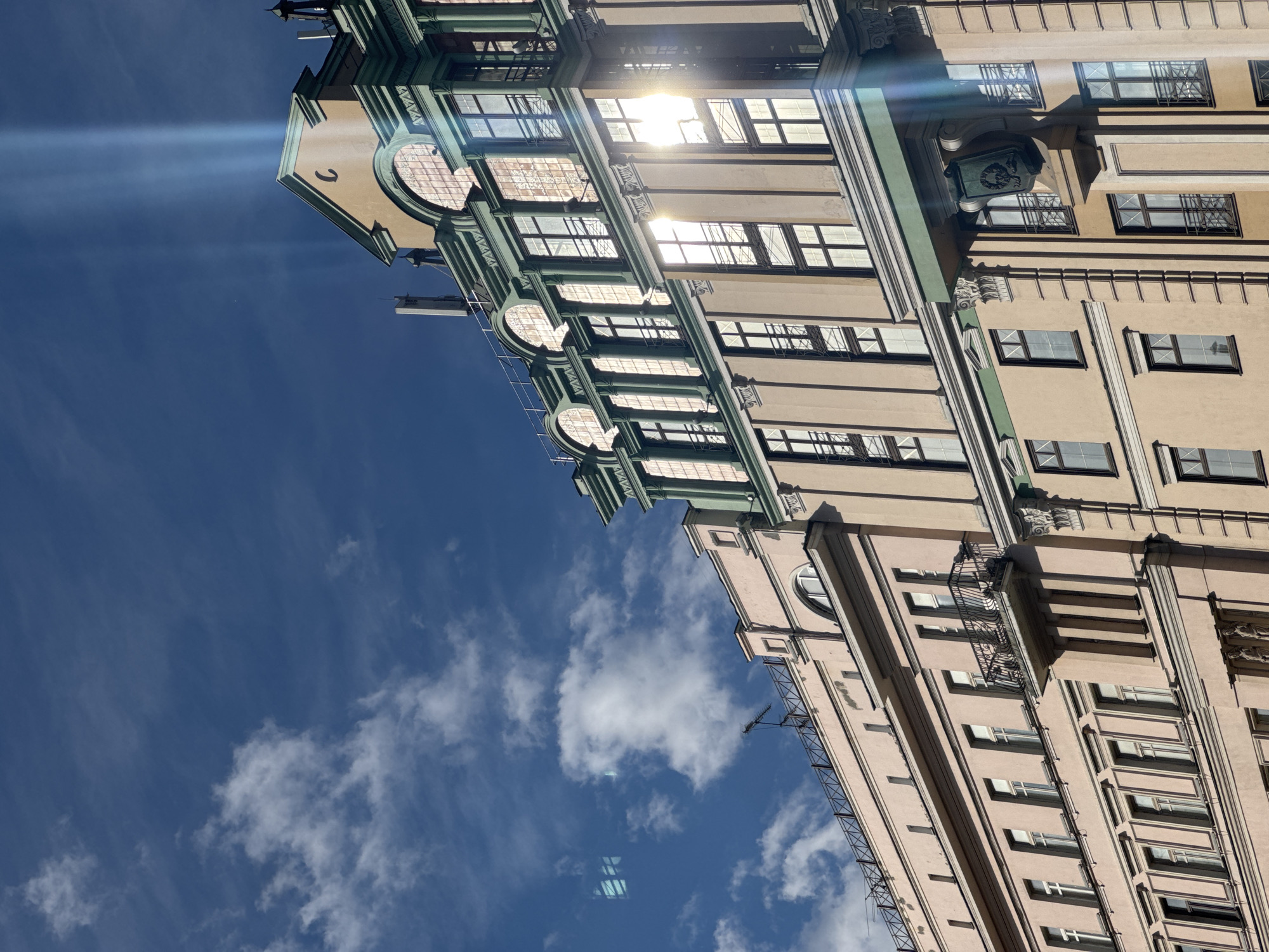 The image shows two buildings in Moscow, Russia, photographed from a low angle against a blue sky with scattered white clouds. The building on the left features beige walls, green trim, and round, decorative windows. The building on the right has light pink walls, windows with dark frames, and ornamental detailing. Sunlight reflects off the windows of both buildings. A metal fire escape is visible on the right building.