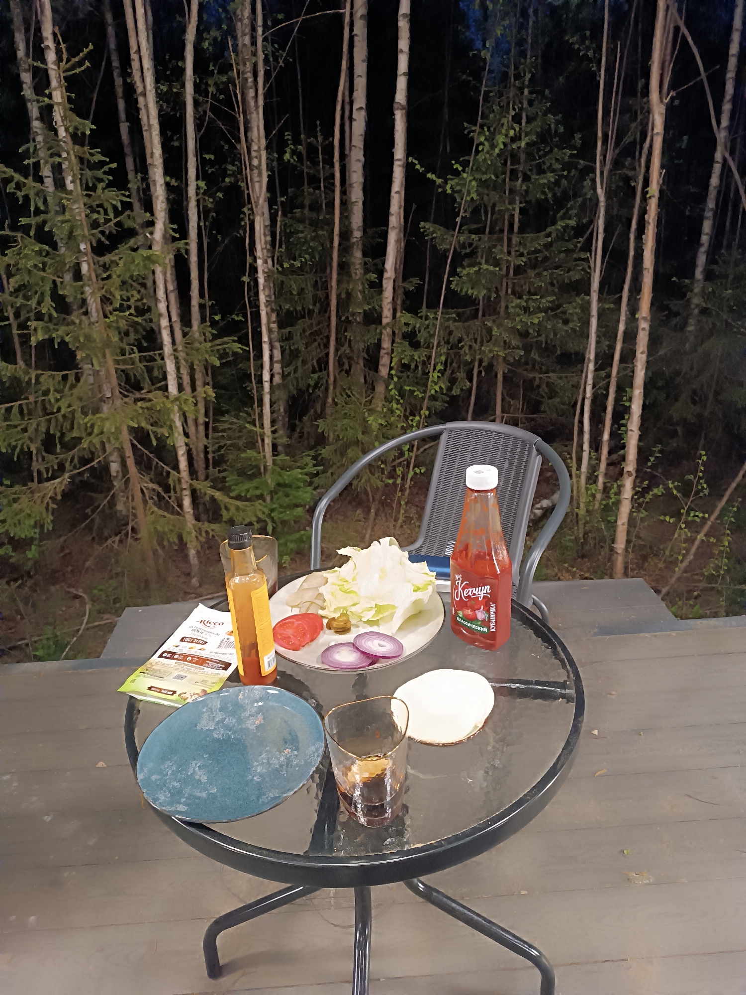 A round, glass-topped table with black metal legs and frame is set outdoors, likely on a deck or patio, with a backdrop of a dense forest visible in the dark. The table displays a partially prepared meal. A plate holds lettuce, sliced tomatoes, and red onion rings. A bottle of salad dressing and a bottle of ketchup are also on the table. Additional items include a blue plate, a glass filled with a dark liquid, a round white object (possibly a bread or tortilla), and a menu. A metal chair with a gray mesh back is positioned behind the table. The setting appears to be in Сепыч, Russia, as indicated by the Cyrillic characters on the ketchup bottle.