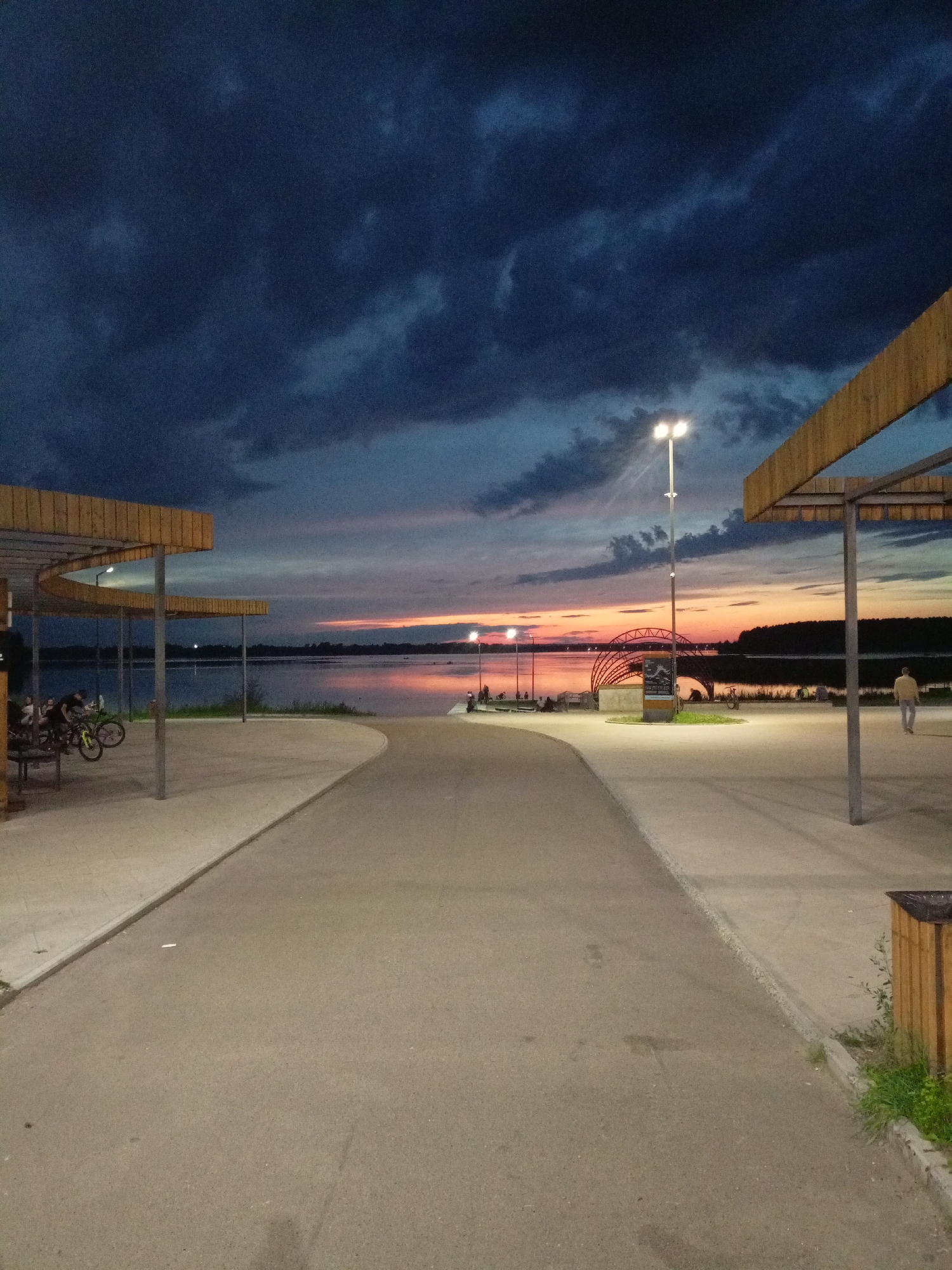 The image depicts a twilight scene at a waterfront promenade in Zheleznogorsk, Russia. A paved pathway curves towards a wide body of water, reflecting the warm hues of the setting sun. Dark, dramatic clouds dominate the upper sky.
On the left, a modern, curved wooden and metal structure provides shade, with several people visible sitting or standing beneath it. Two bicycles are parked near this structure.
On the right, another similar wooden and metal shelter is partially visible. A single individual is walking away from the camera on the paved promenade. In the distance, a large, arched metal structure is illuminated. The shoreline on the opposite side of the water is lined with trees. The overall atmosphere is calm and serene, with artificial lighting from streetlamps beginning to illuminate the area.