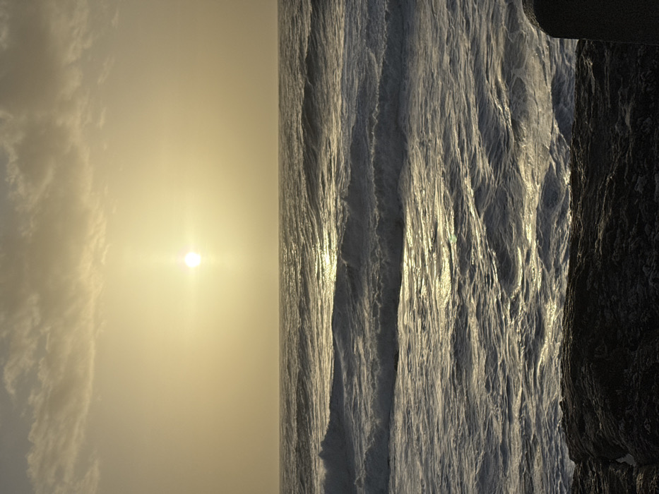 The image depicts an ocean landscape at sunset. The sky is a hazy yellow with a visible sun emitting bright light. Ripples cover the ocean surface, reflecting the sunlight in shimmering patterns. To the right, dark, textured rocks border the ocean. The photograph is rotated, presenting the sky on the left and the water vertically oriented.